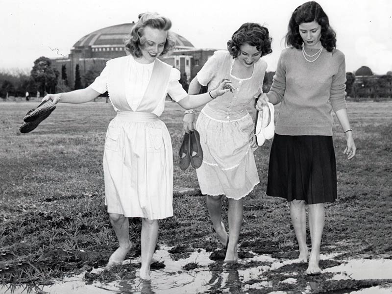 Three Newcomb College students pick their way across a soggy field in front of McAlister Auditorium,circa 1950. 