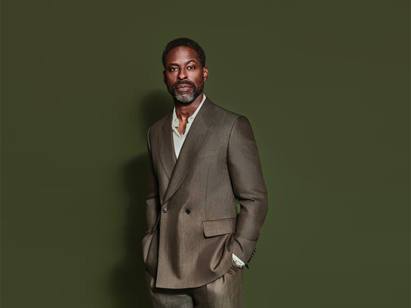 Sterling K. Brown in brown double-breasted suit, hands in pockets, against a dark green background. 