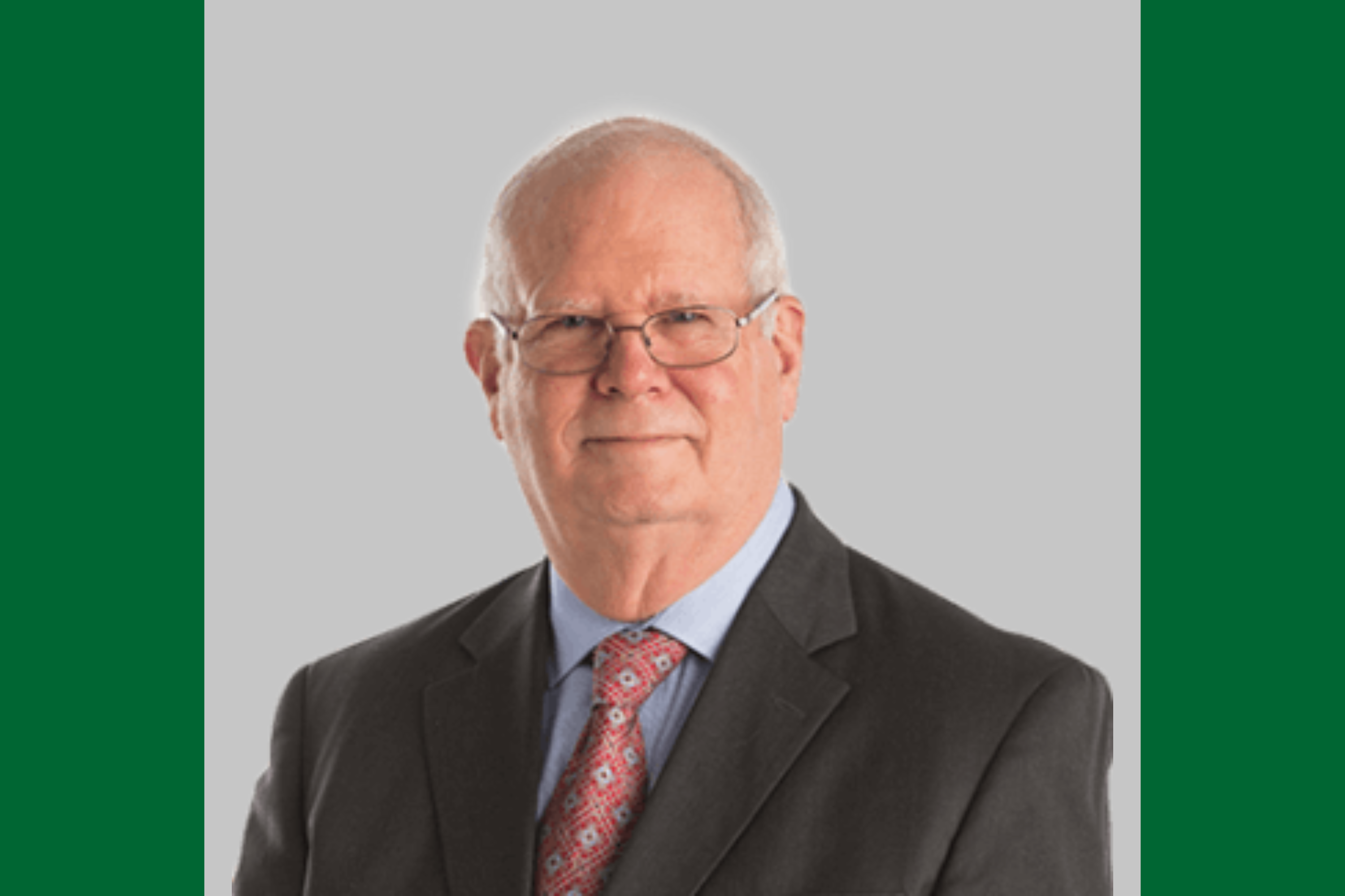 Older man in suit, tie, and glasses, smiling in a professional photograph. 