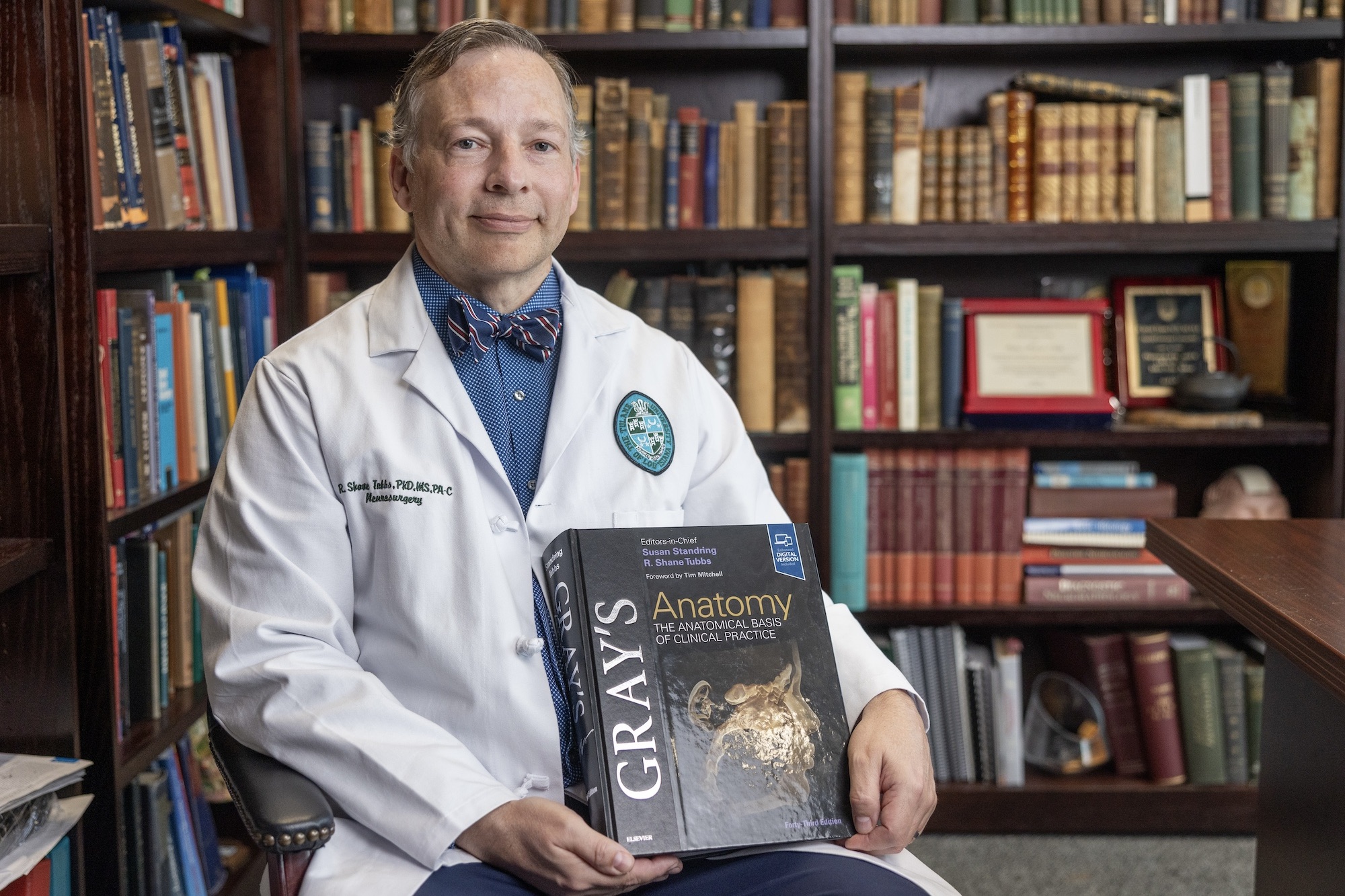 Dr. Shane Tubbs in lab coat and bow tie smiles, holding Gray&amp;#039;s Anatomy book before a bookshelf. 