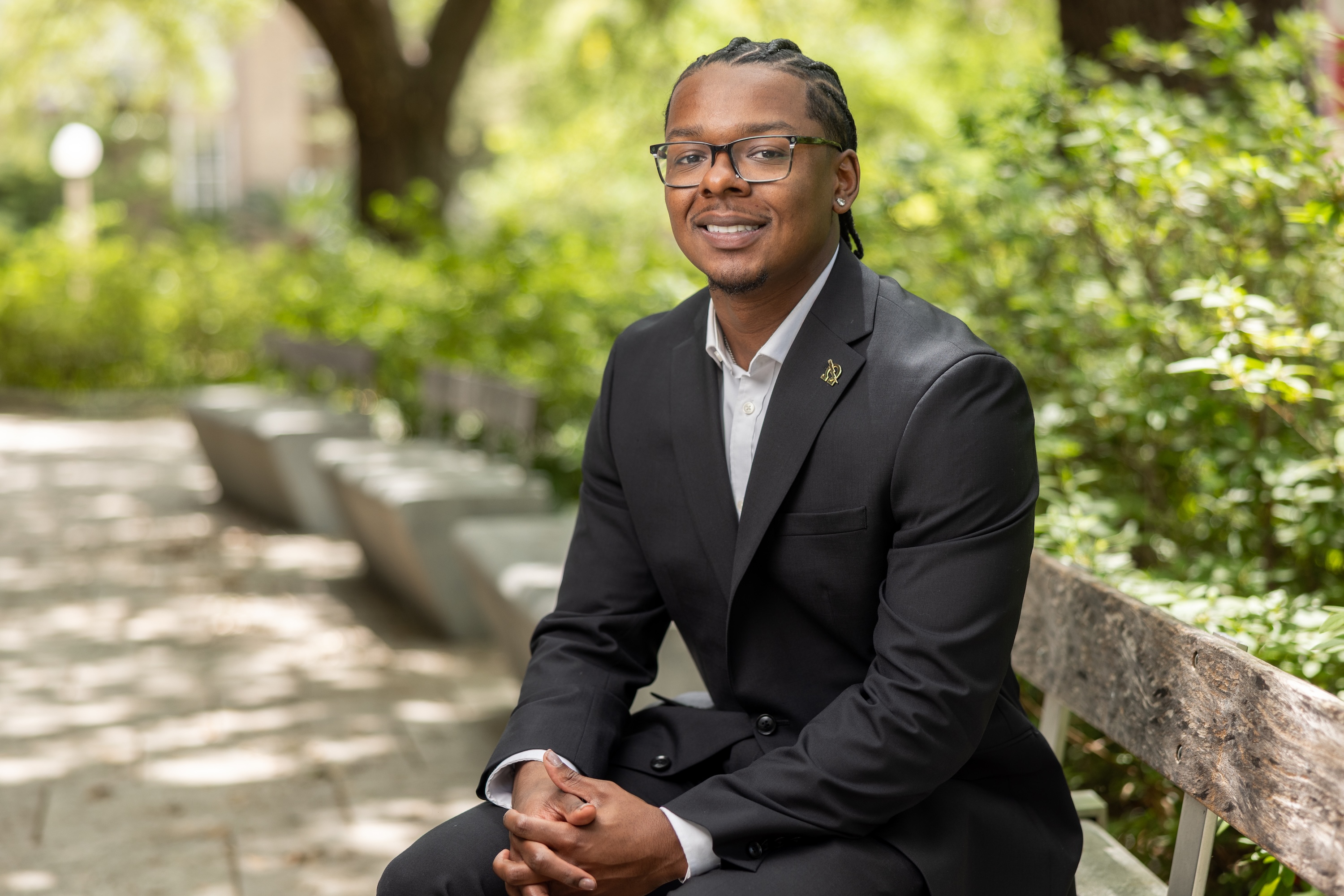 Rodrek in a black suit and glasses sits on an outdoor bench. 