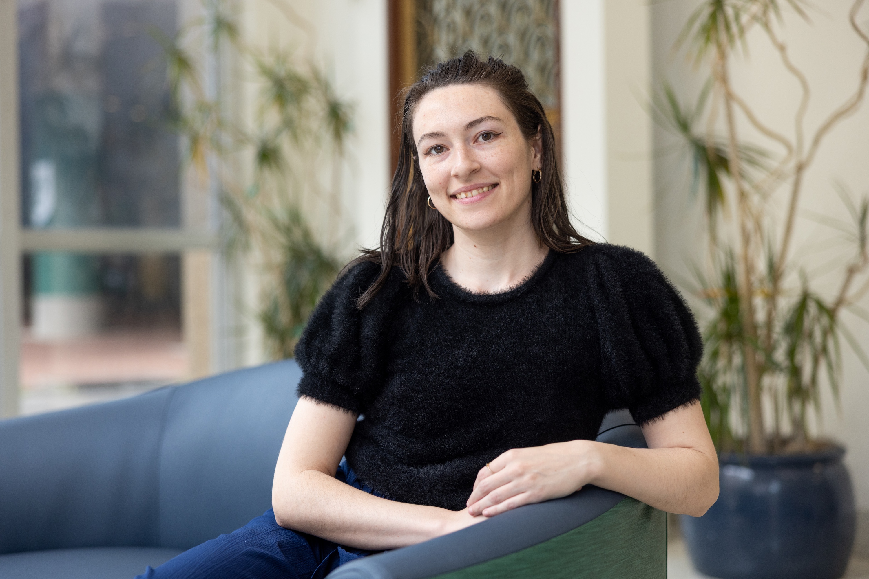 
Smiling woman with dark hair, wearing a black fluffy top, sitting on a blue sofa. 