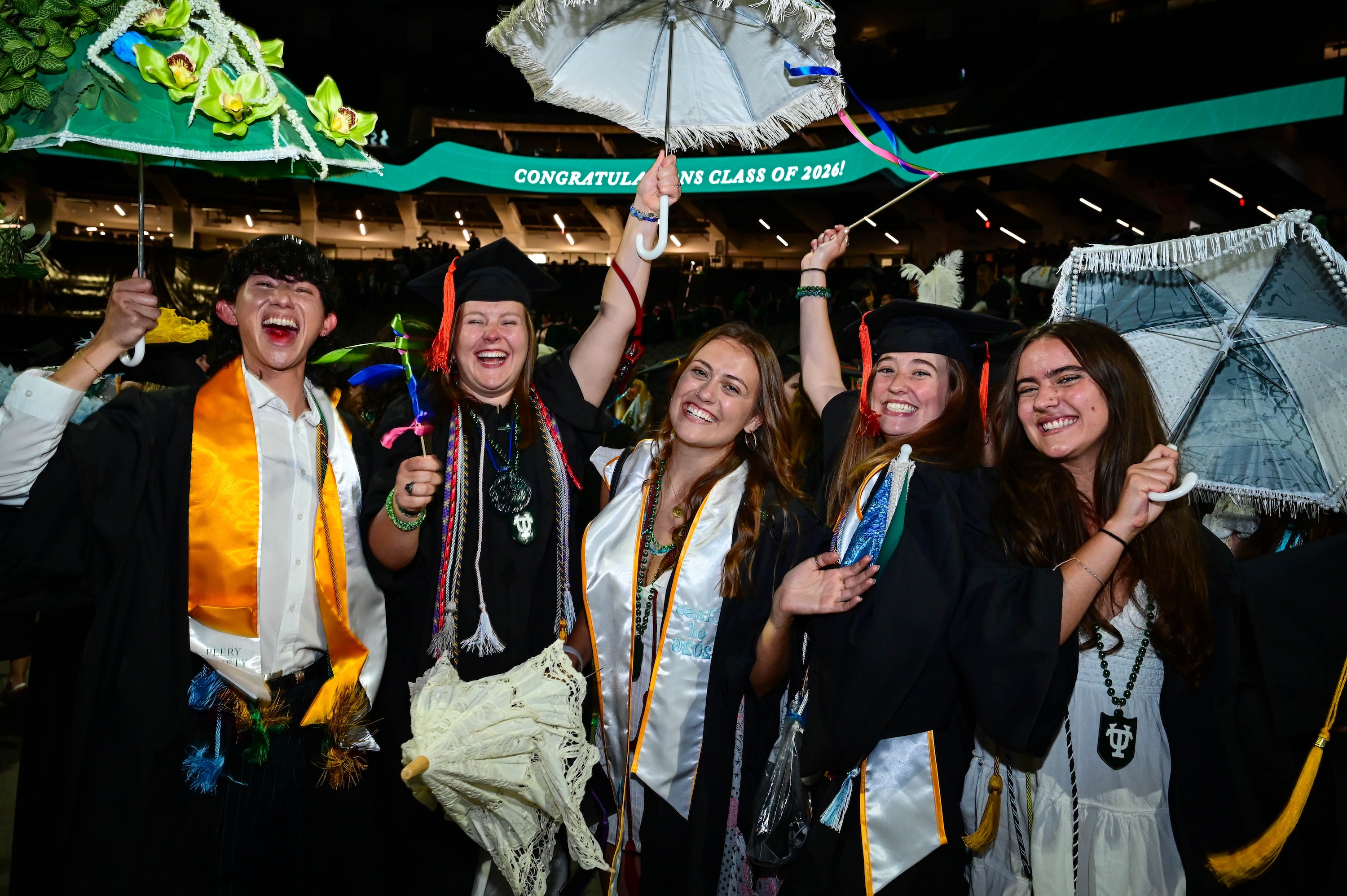 
Five joyful graduates from the Class of 2026 hold umbrellas, celebrating. 