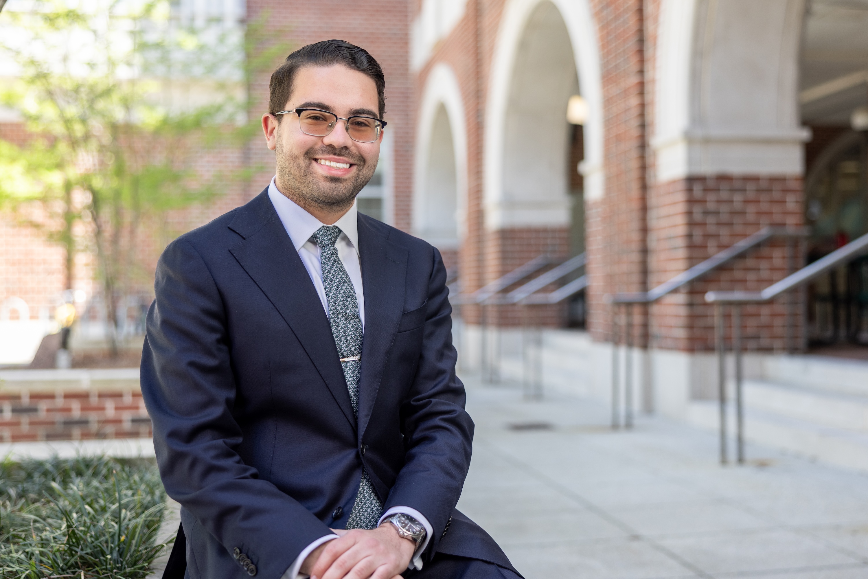 
Smiling man in a dark suit and glasses sitting outdoors near a brick building. 