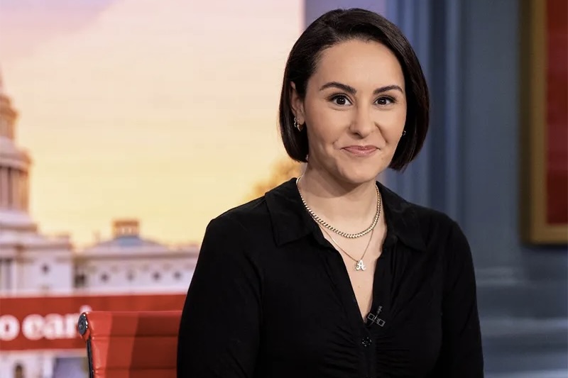 
Young woman with dark hair, black top, smiling in a TV studio with government building background. 