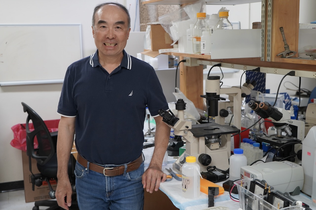 
YiPing Chen blue polo shirt smiles in a laboratory with a microscope. 
