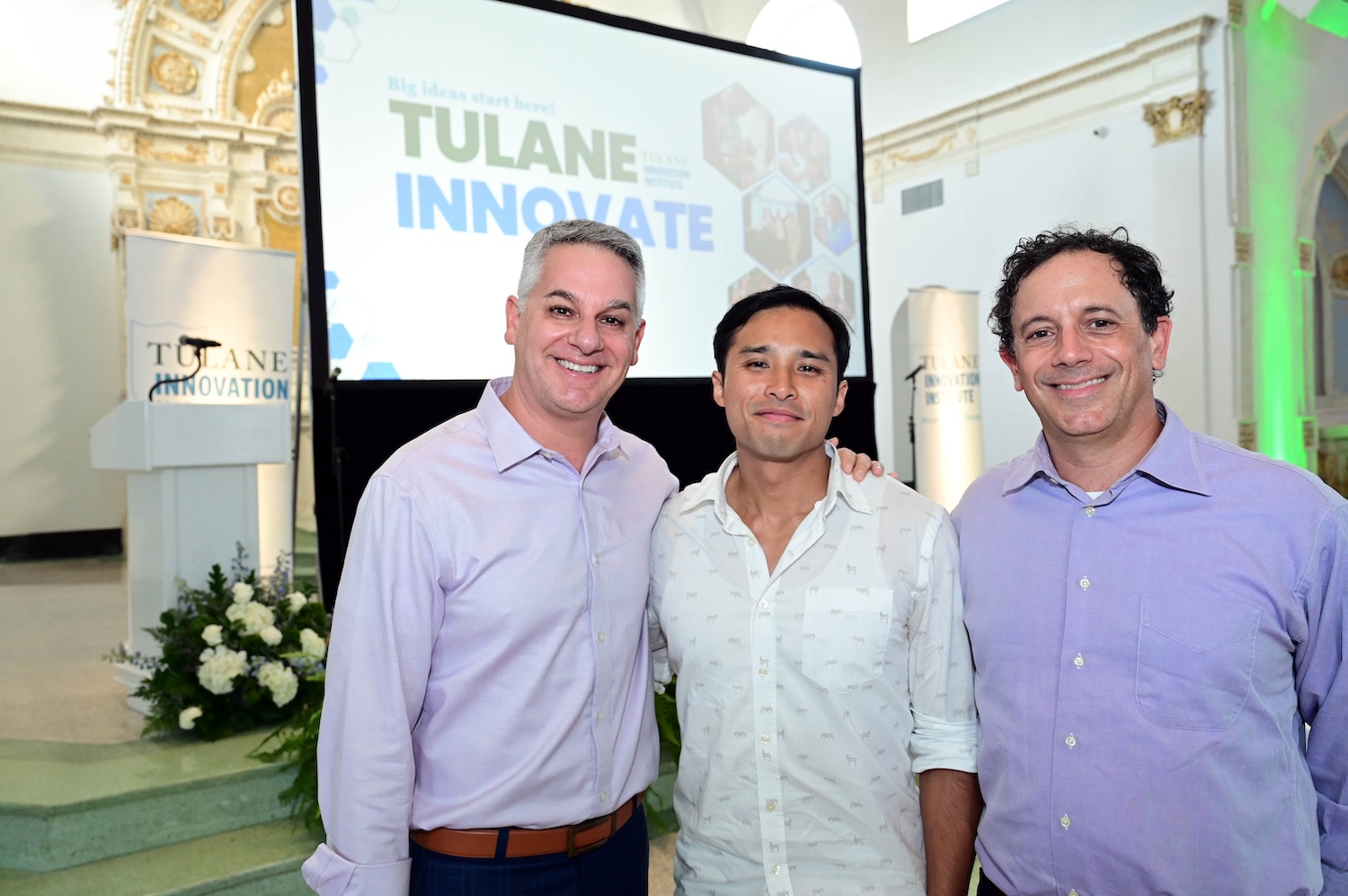 
Three smiling men pose at a Tulane Innovate event, screen visible in background. 