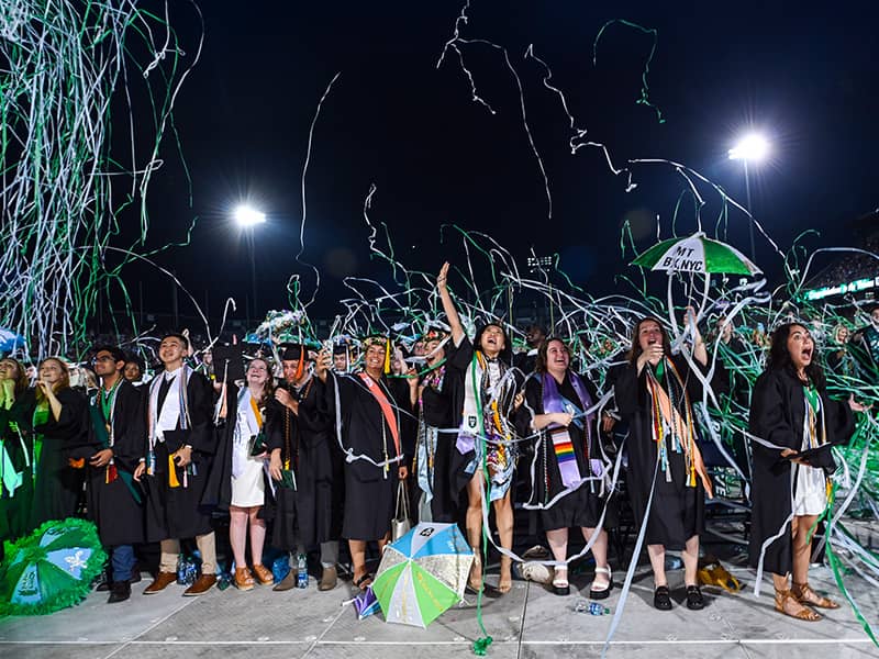 Elated students celebrate their Commencement as olive and blue streamers surround them. 