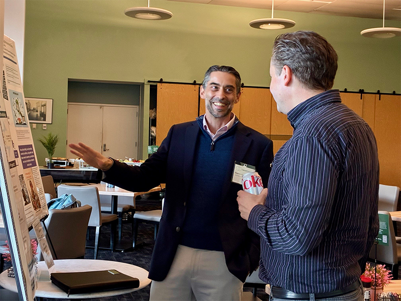 Two men stand in conversation beside a research poster in a conference room.