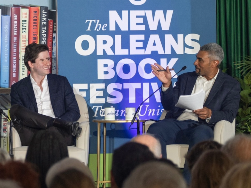Two men on stage, one smiling, the other gesturing, with a New Orleans Book Festival backdrop.