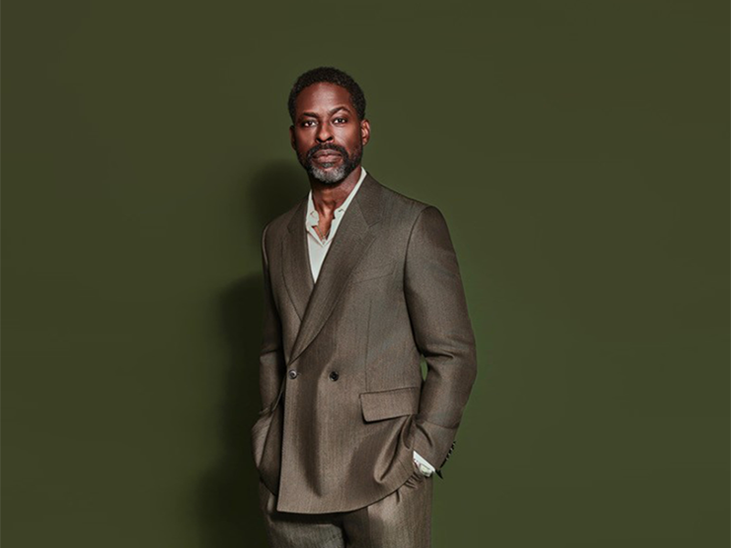 Sterling K. Brown in brown double-breasted suit, hands in pockets, against a dark green background.
