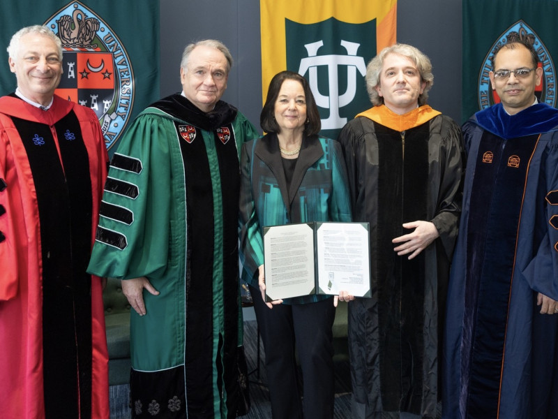 Four people in academic regalia and one woman in a suit, holding a document, pose by banners.