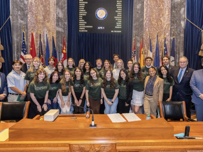 Group of young people in green shirts with adults in suits pose in a formal chamber.