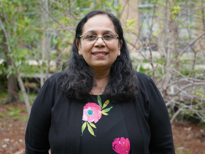 Smiling woman with dark hair and glasses wearing a black floral top, green foliage background.