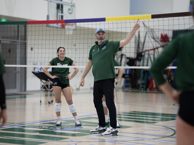 Volleyball coach in a green shirt gestures to female players on an indoor court.