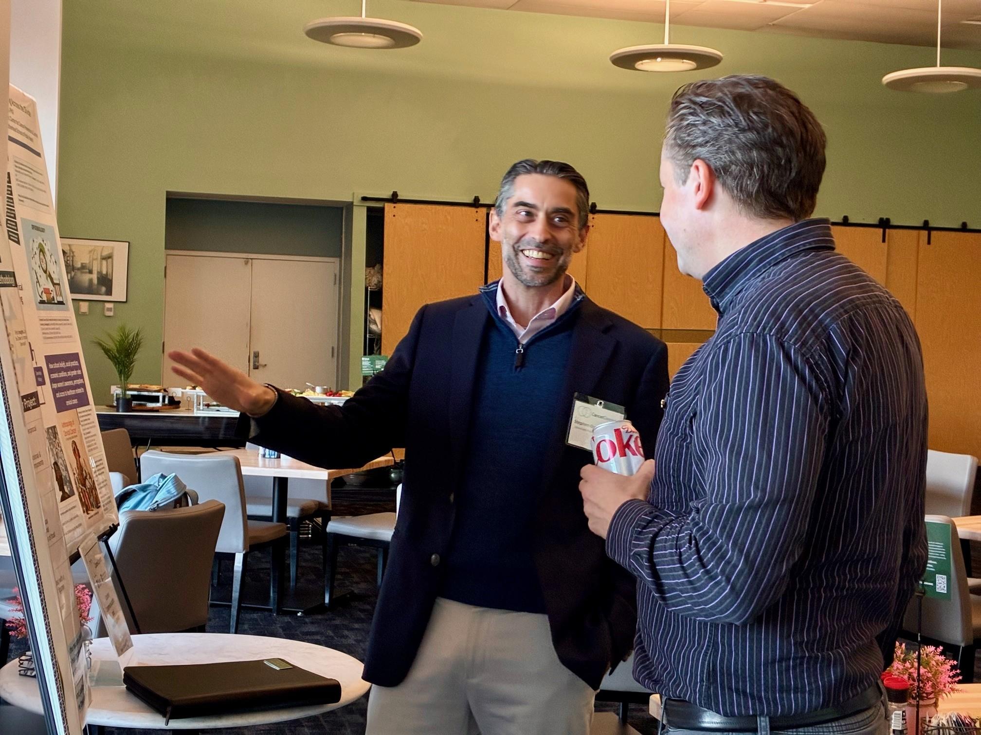 Two men stand in conversation beside a research poster in a conference room.