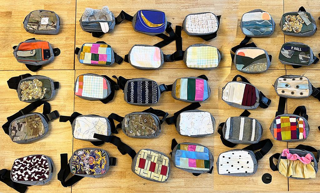 Dozens of small, handmade fanny packs arranged in neat rows on a wooden floor, each featuring different colorful fabrics, patterns, and textures.