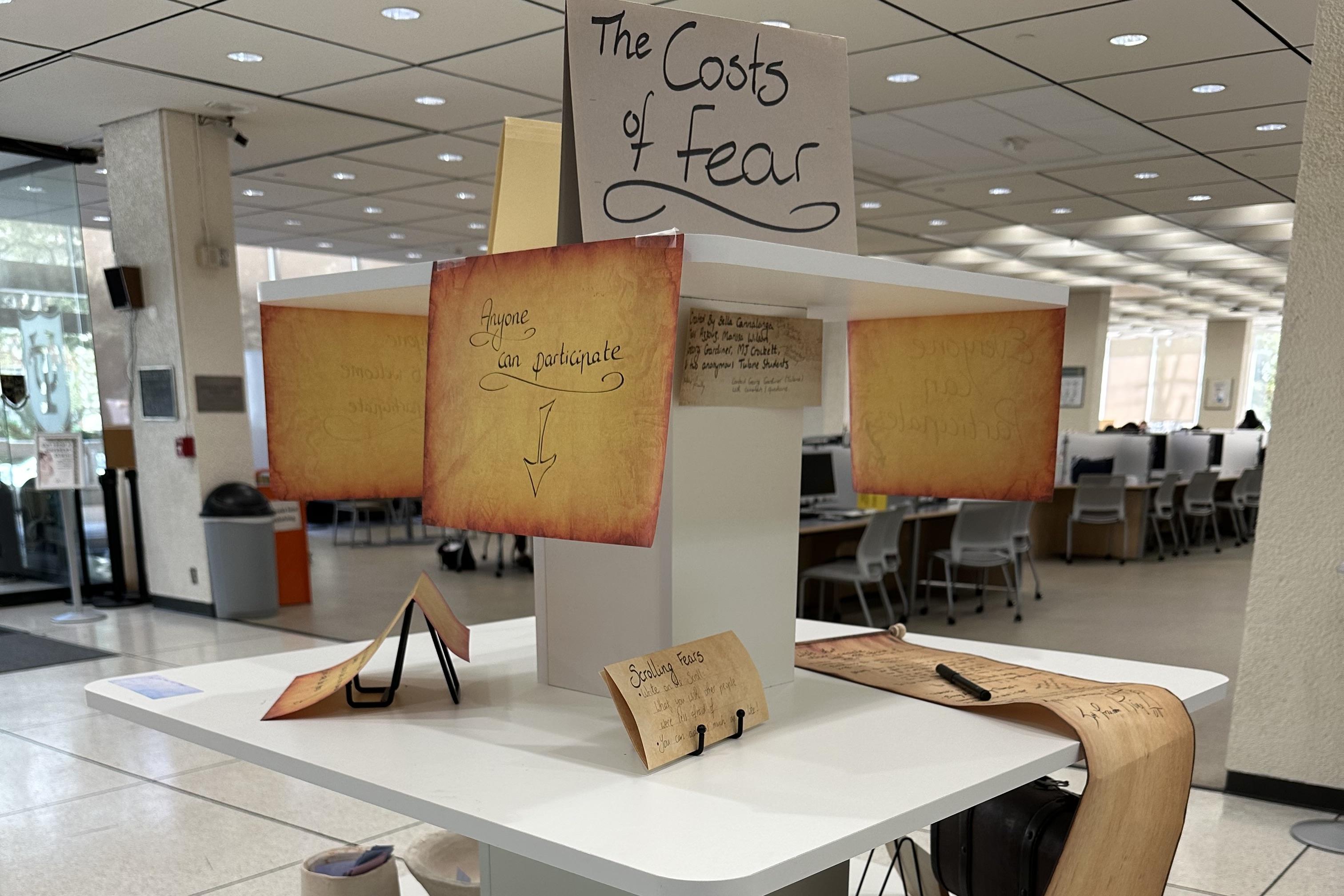 Library display titled "The Costs of Fear" with scroll-like papers on a white stand.