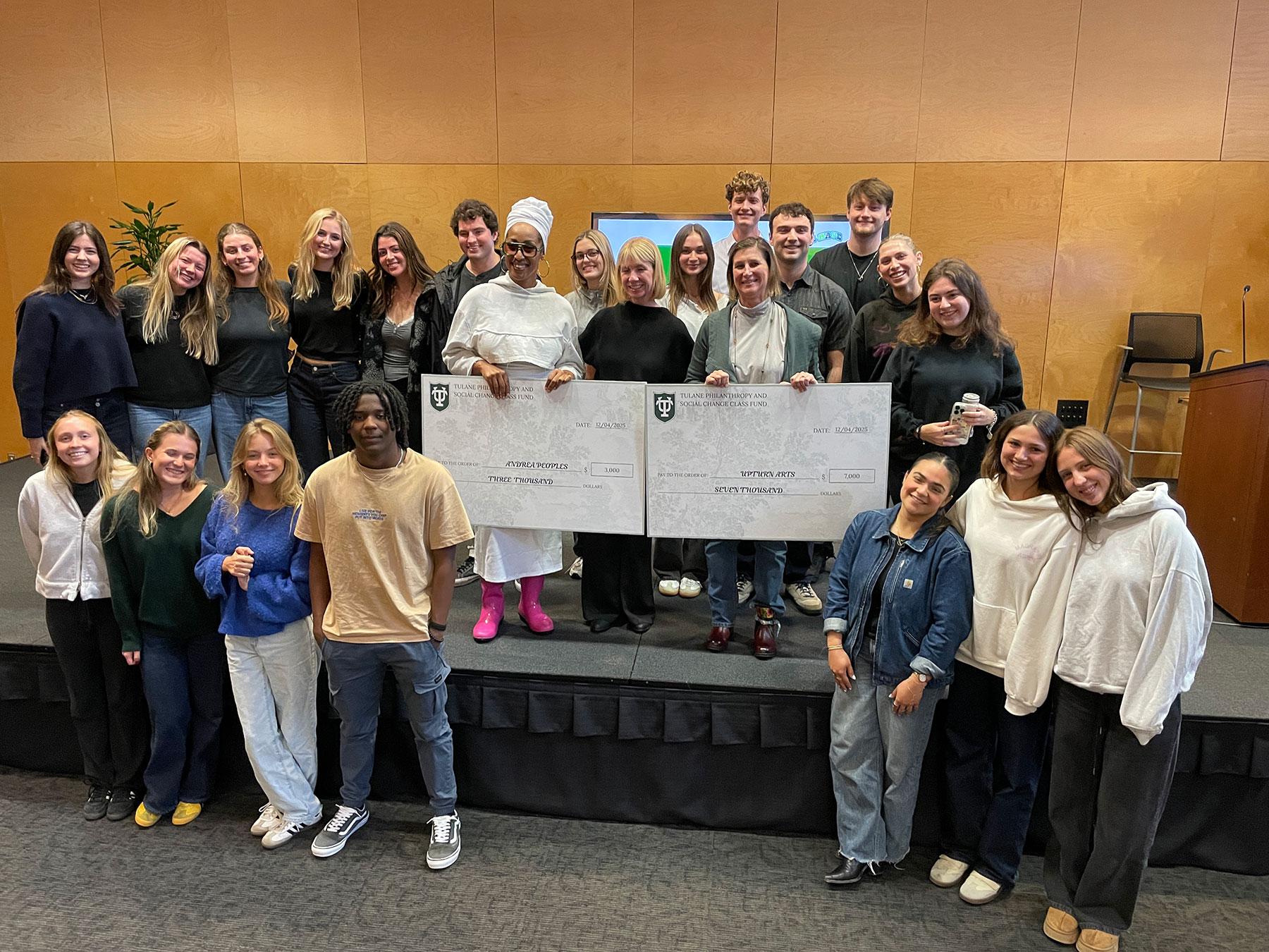 Smiling group of students and adults holding two oversized checks on a stage.