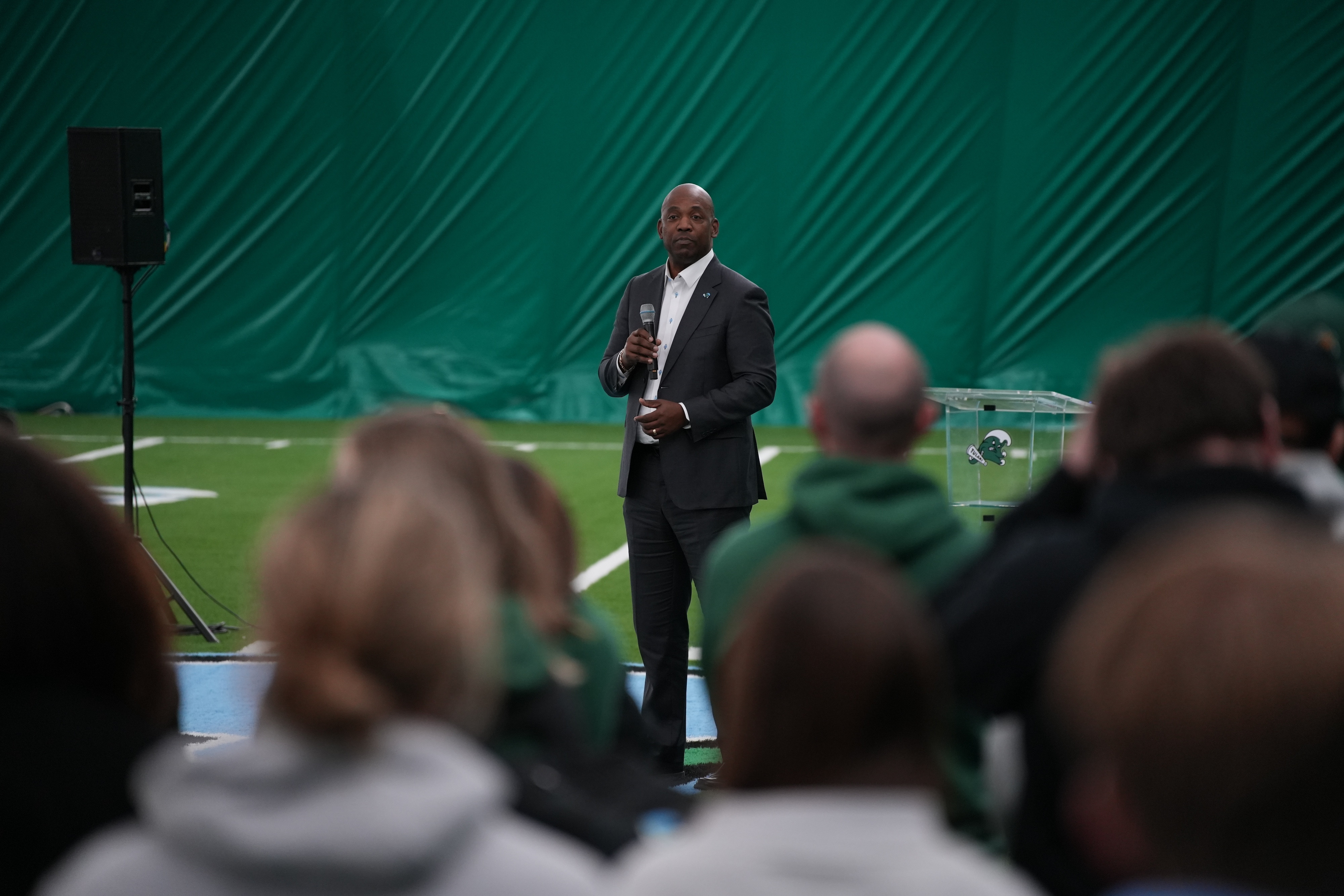 David Harris in dark suit speaks into microphone to audience on an indoor green field.