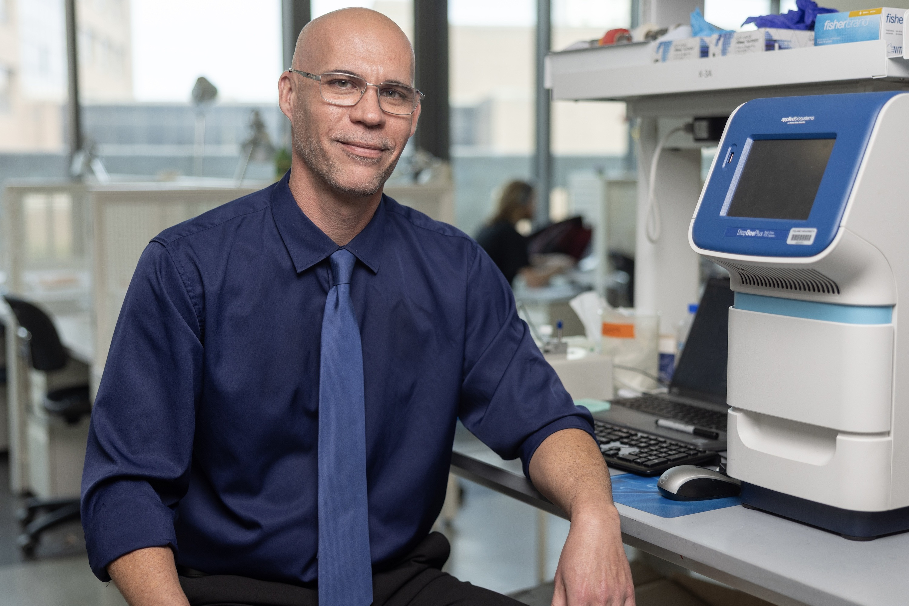 Mark Mondrinos in glasses, blue shirt, and tie smiles in a laboratory with equipment.