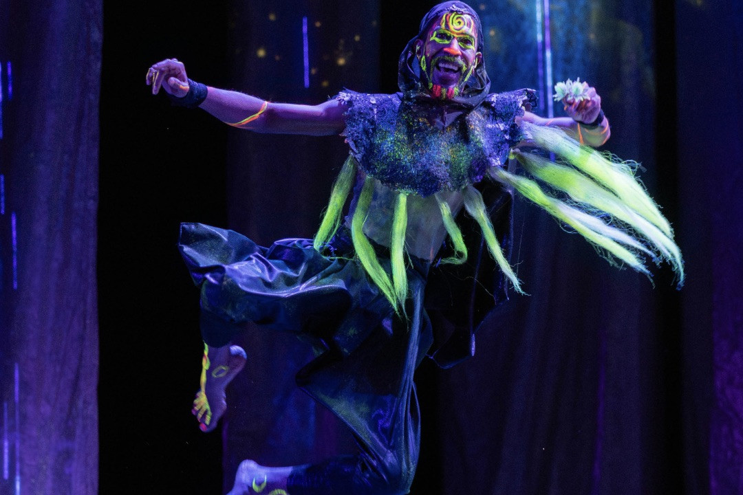 Acrobat with vibrant UV body paint and costume mid-jump against a dark background.