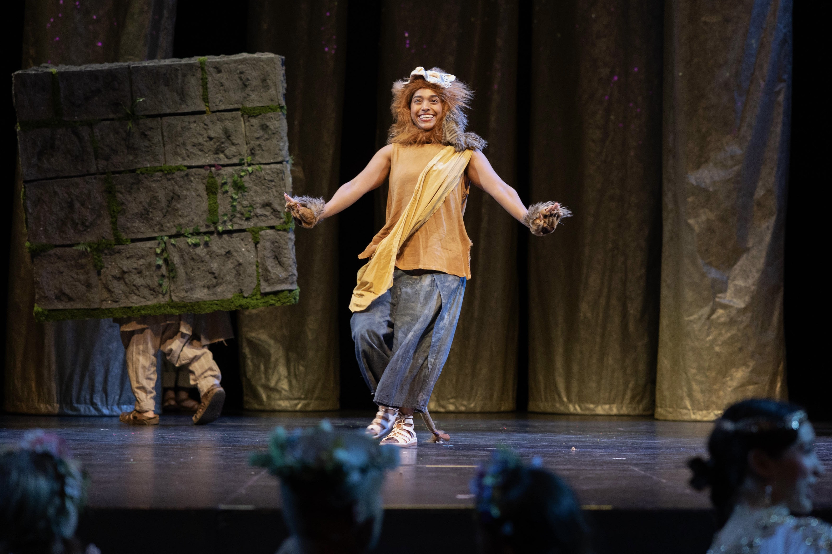 Smiling person in lion costume on stage, dancing near a large gray block.