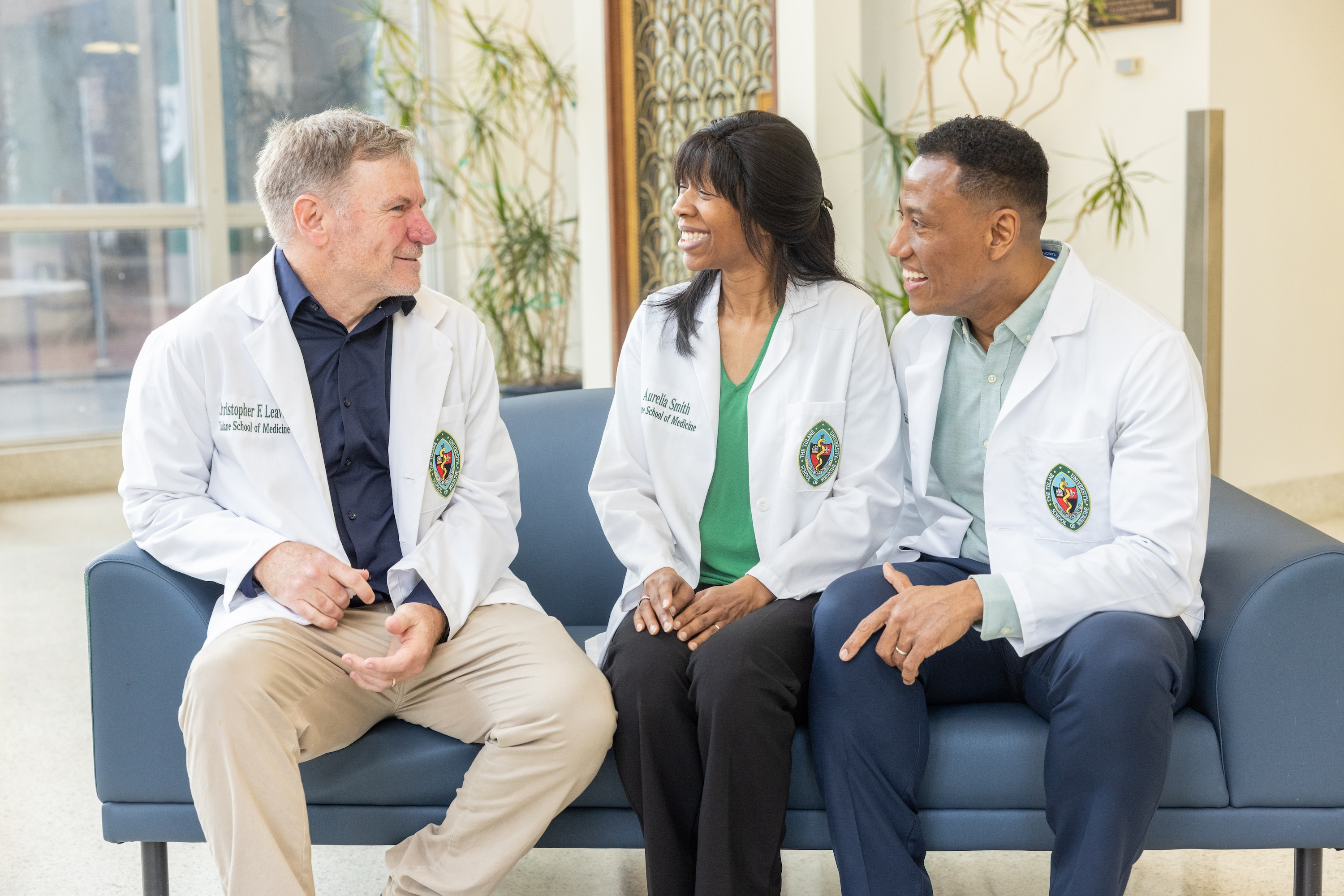 Three medical professionals in lab coats sit on a blue couch, smiling and talking.