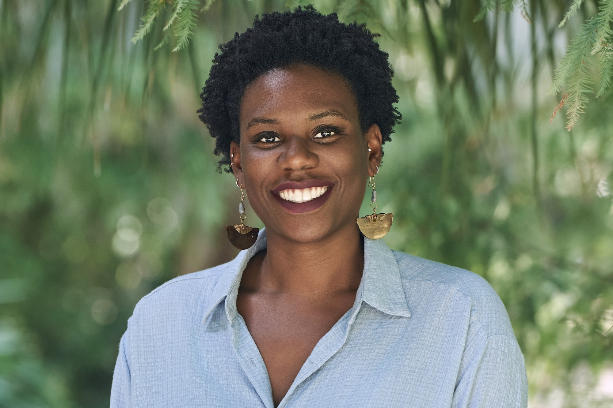 Smiling Black woman with curly hair and gold earrings, light blue shirt, green background.
