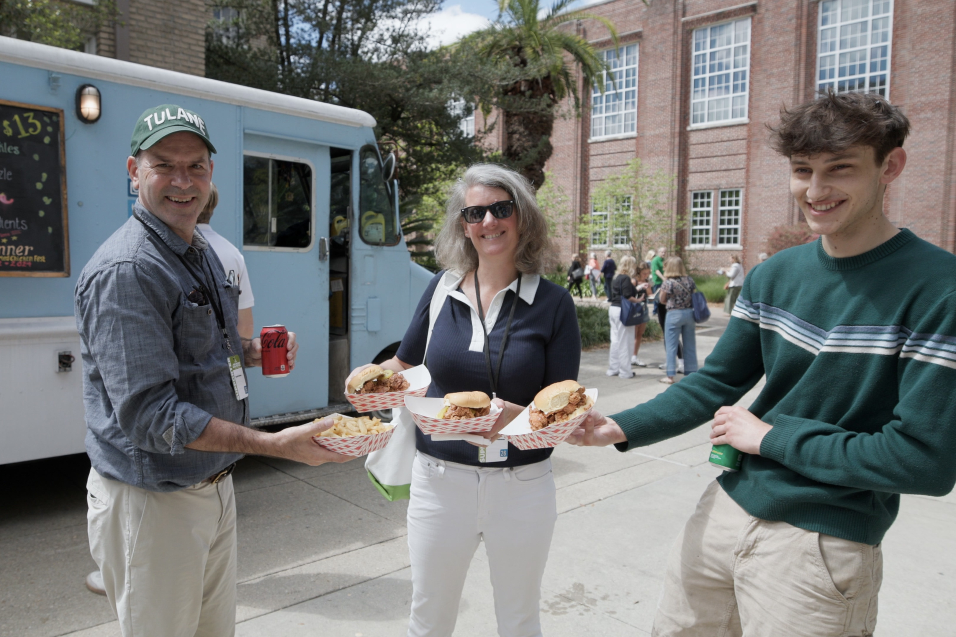 Three smiling people hold burgers and fries from a light blue food truck.