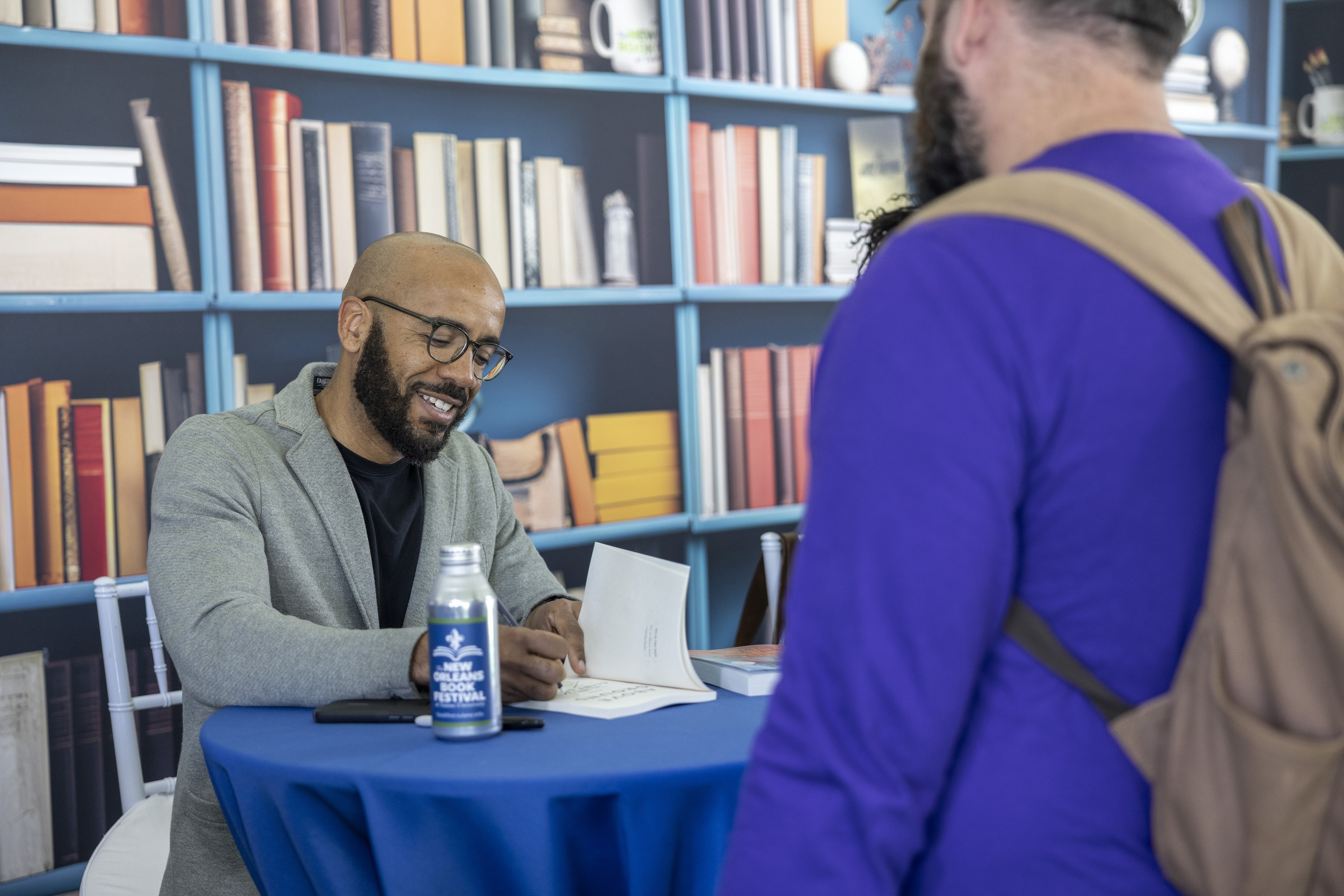 Clint Smith signs at a table, a person with a backpack observes.