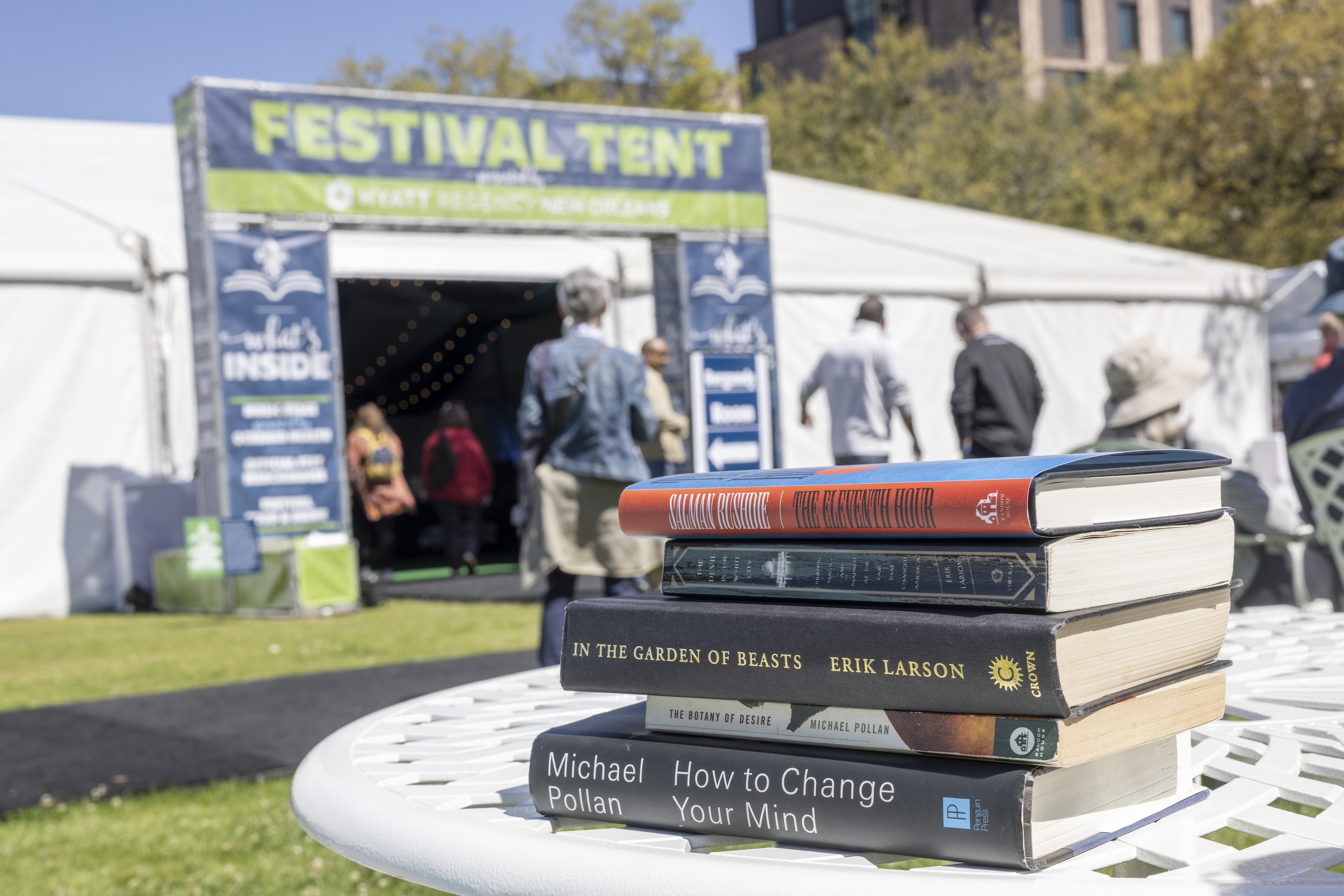 Stack of books on a white table, a festival tent and people in the background.