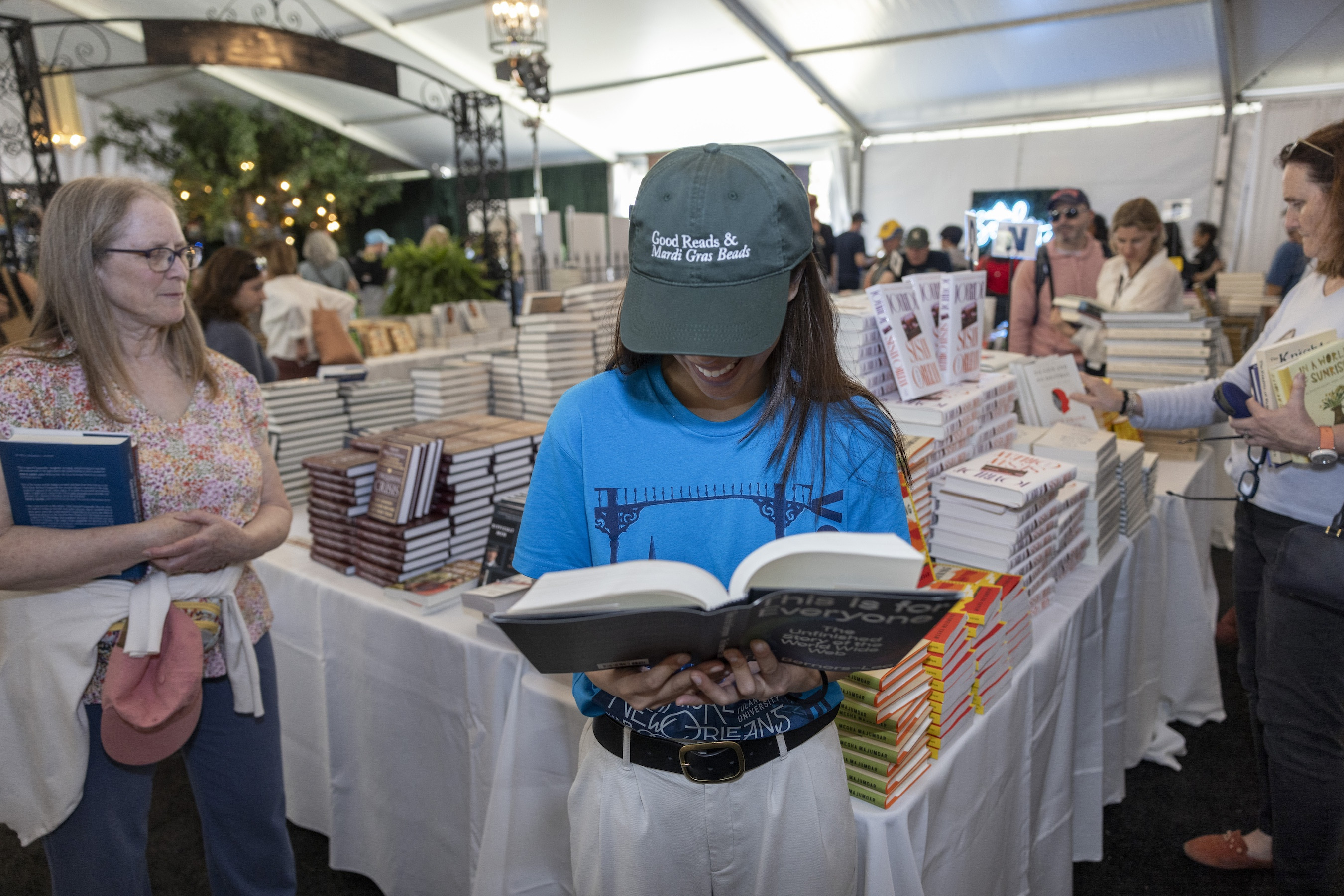 Young person in green hat engrossed in reading a book at a bustling outdoor book fair.