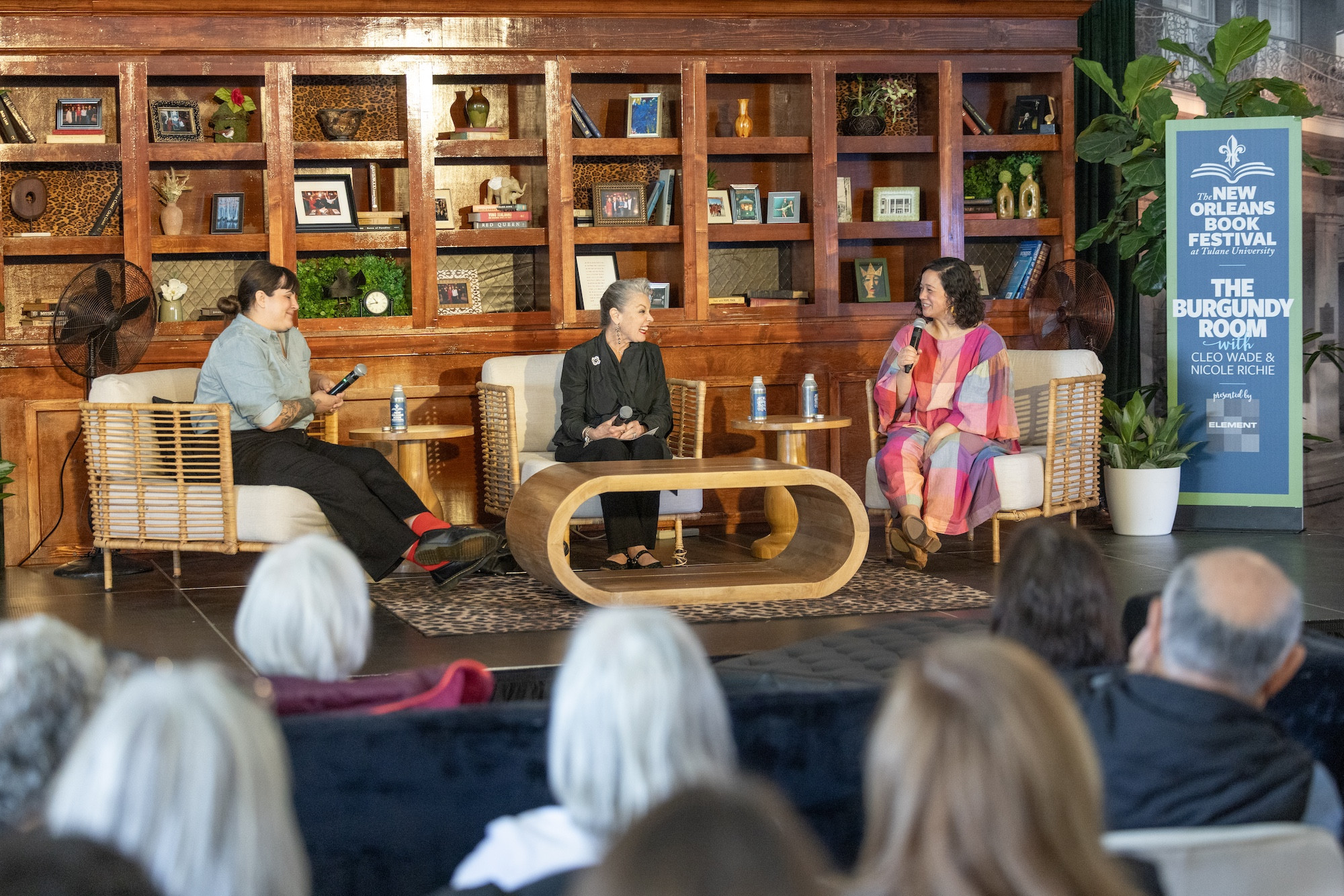 Three women speak on a panel stage with a bookshelf backdrop, facing an audience.