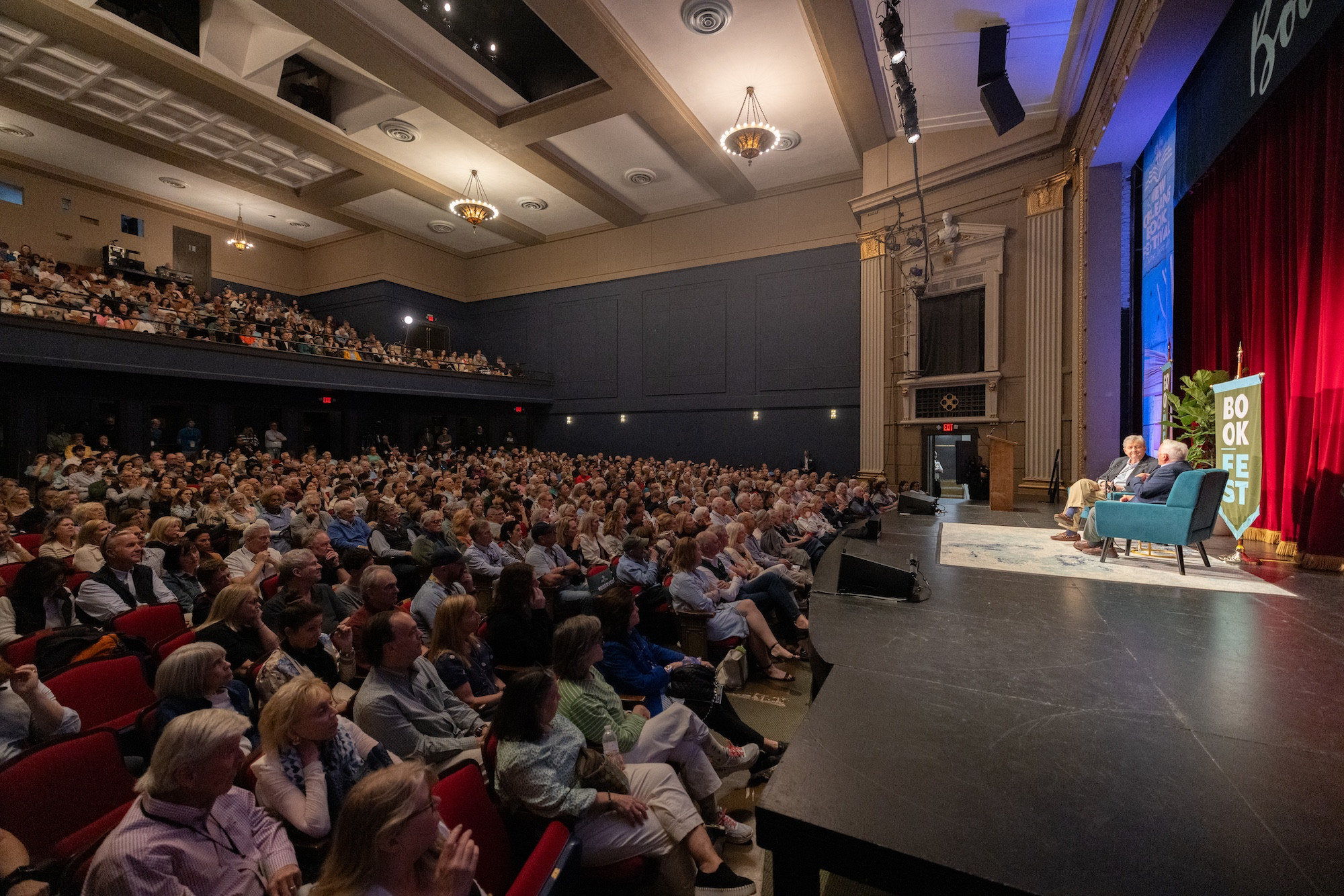 Large audience gathered in a theater watching two speakers on stage with red curtains.