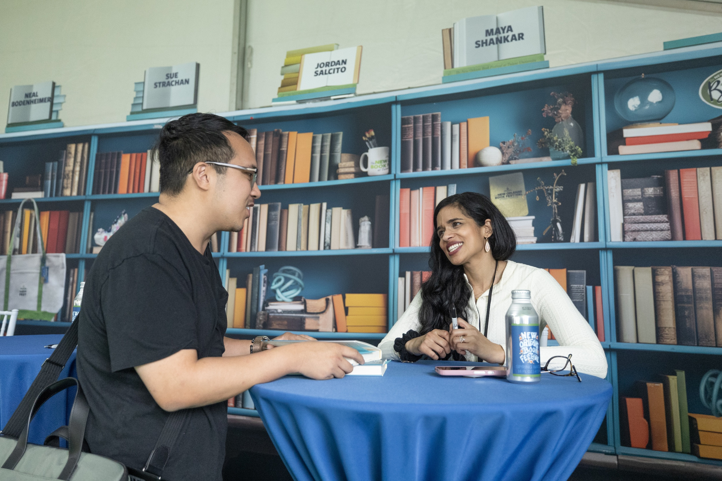 Smiling man and woman talk at a blue table in front of a bookshelf graphic.