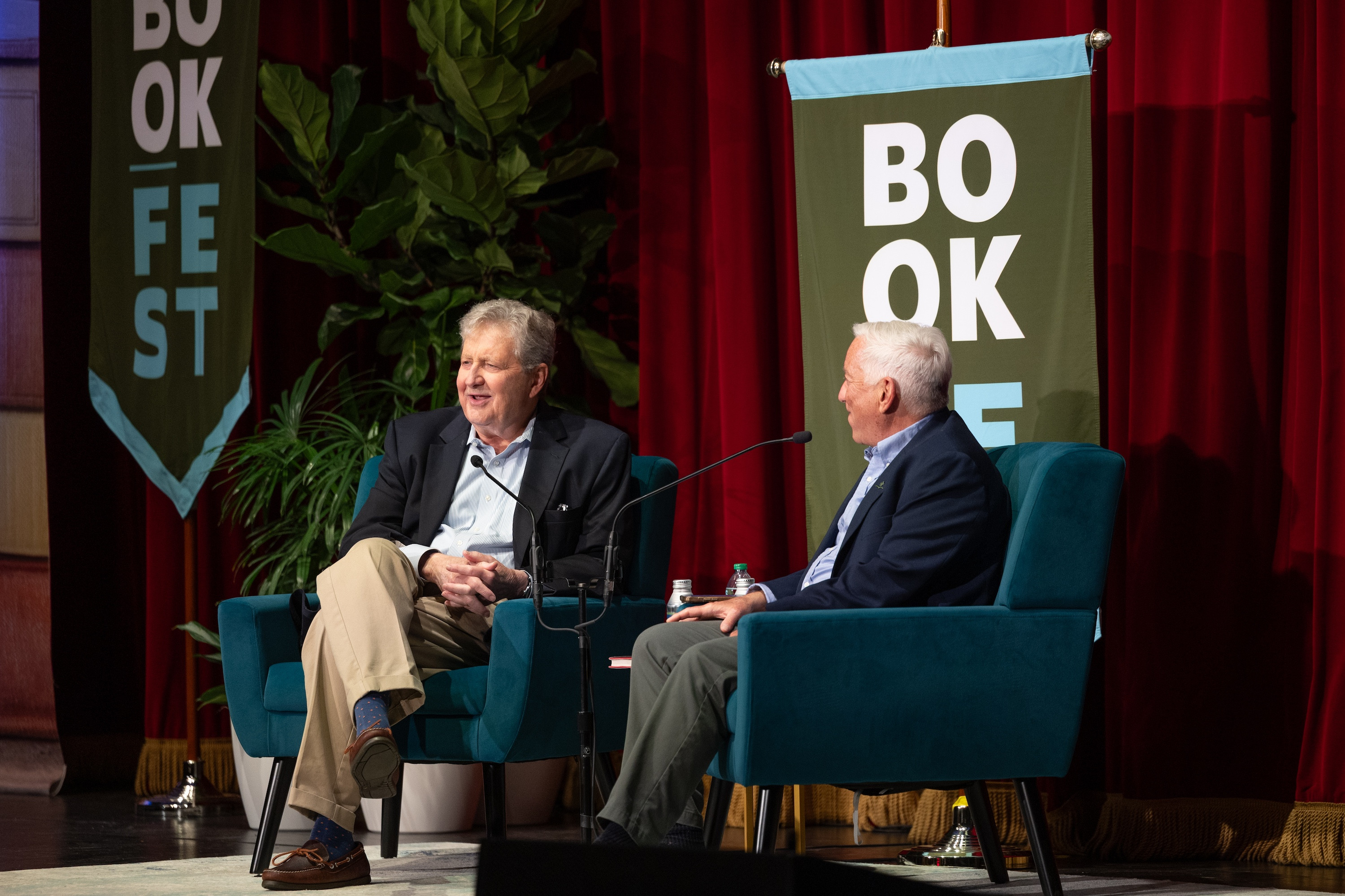 Two smiling men in teal chairs converse on a stage at Bookfest.
