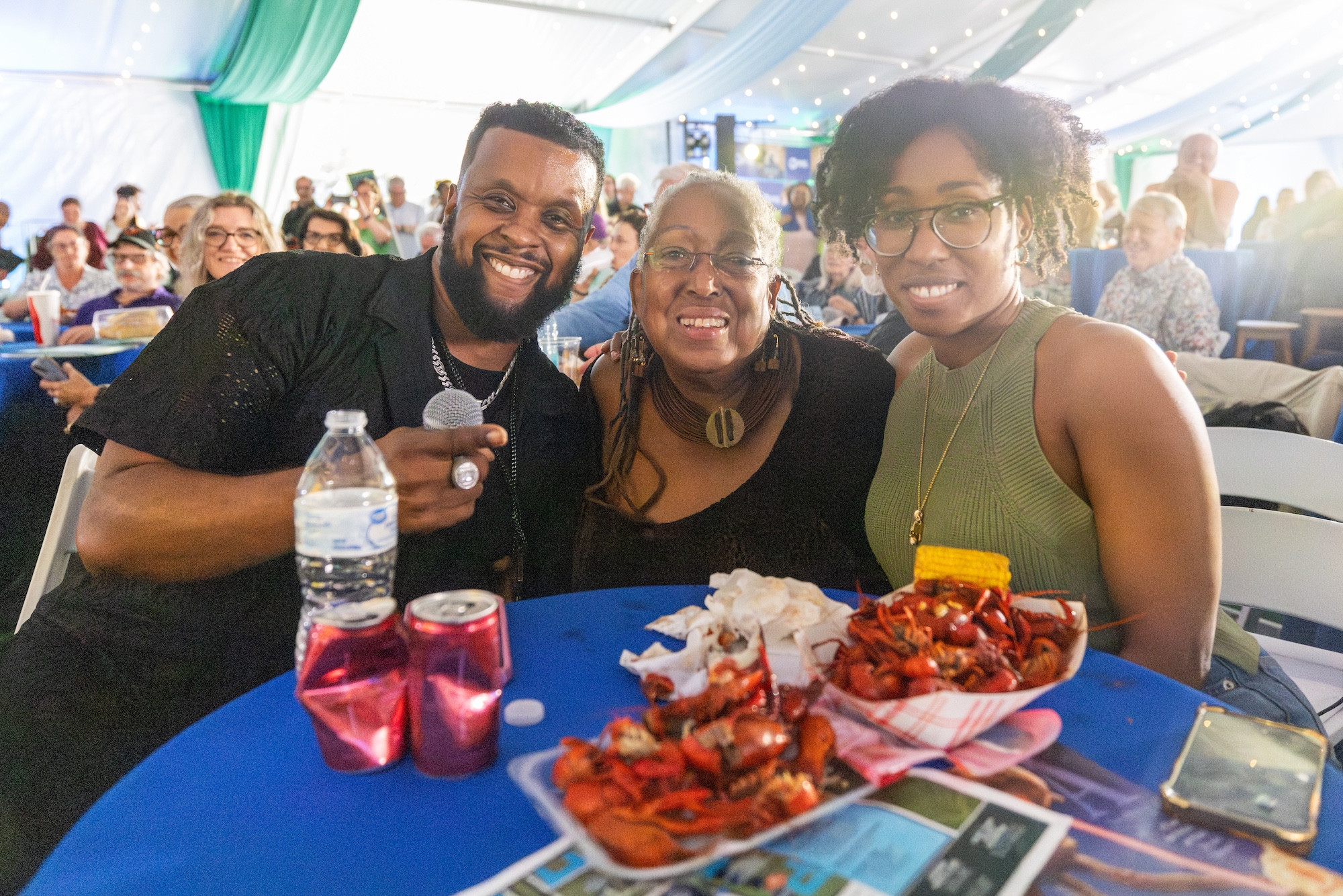 Three smiling people at a table with crawfish and drinks under a tent.