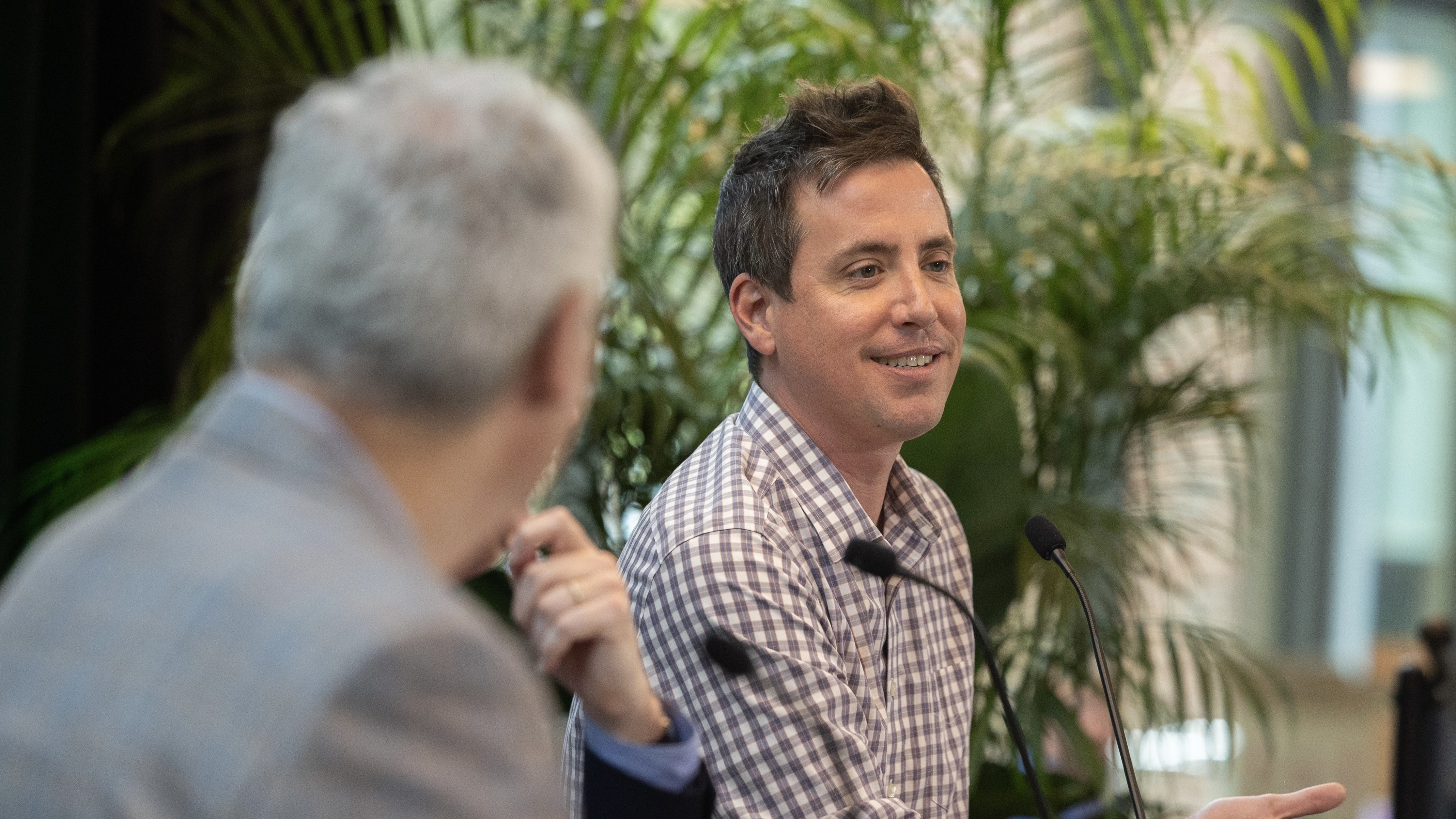 Nicholas Mattei smiles, speaking into a microphone with another person and plants behind