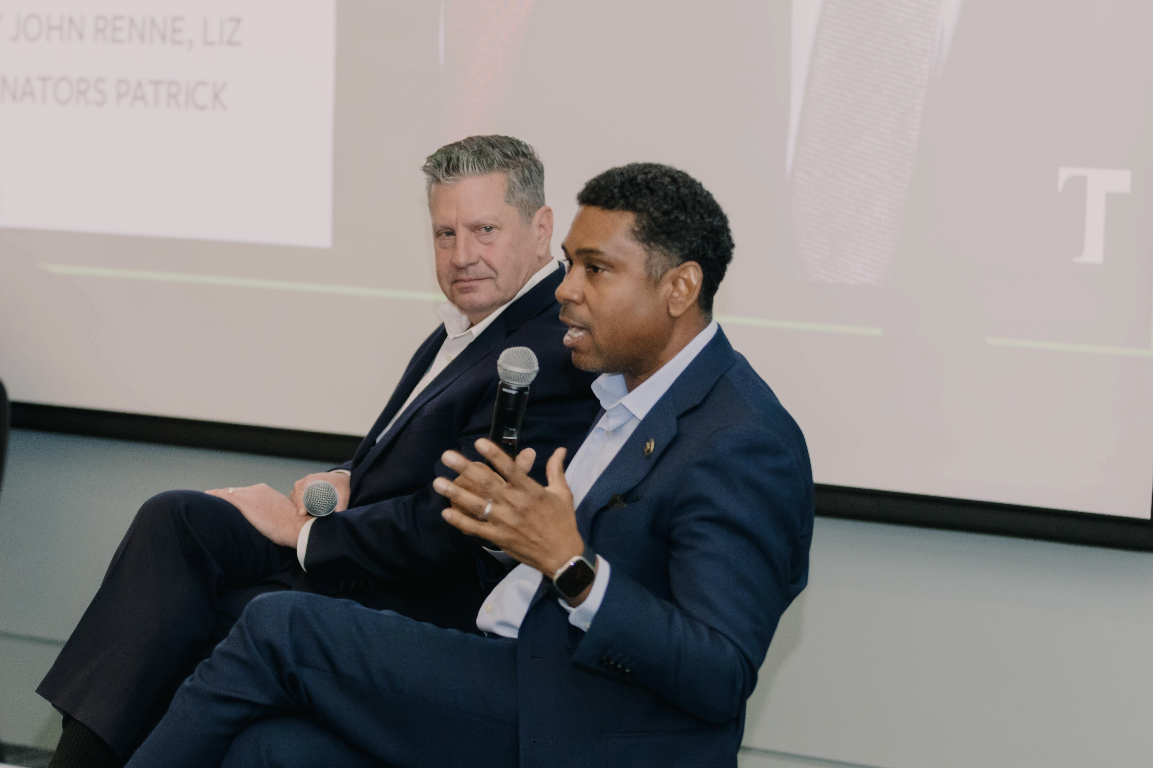 Gary Carter Jr. in blue suit speaking into a microphone, seated beside Patrick Connick listening.