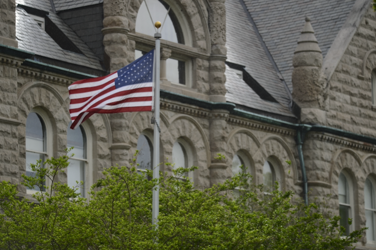 US flag flying on a pole in front of Gibson Hall, a historic stone building with green foliage.