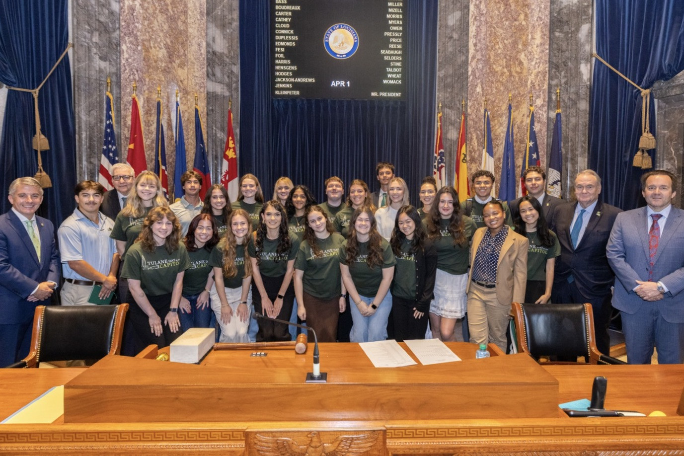 Group of young people in green shirts with adults in suits pose in a formal chamber.
