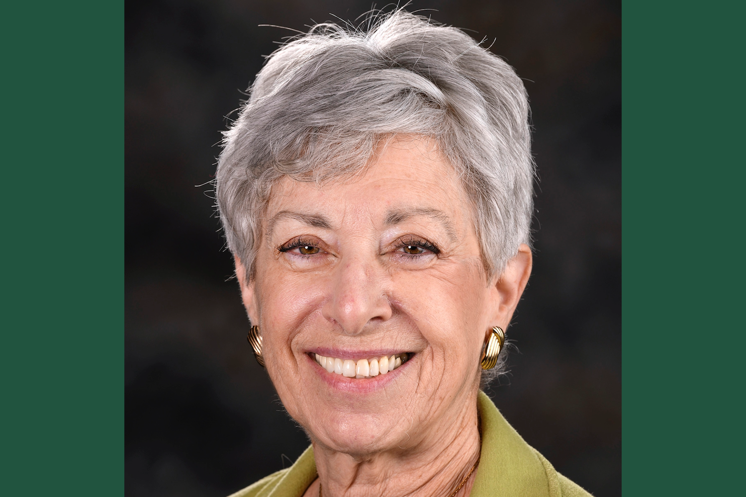 Smiling older woman with short gray hair, wearing a green top and gold earrings.