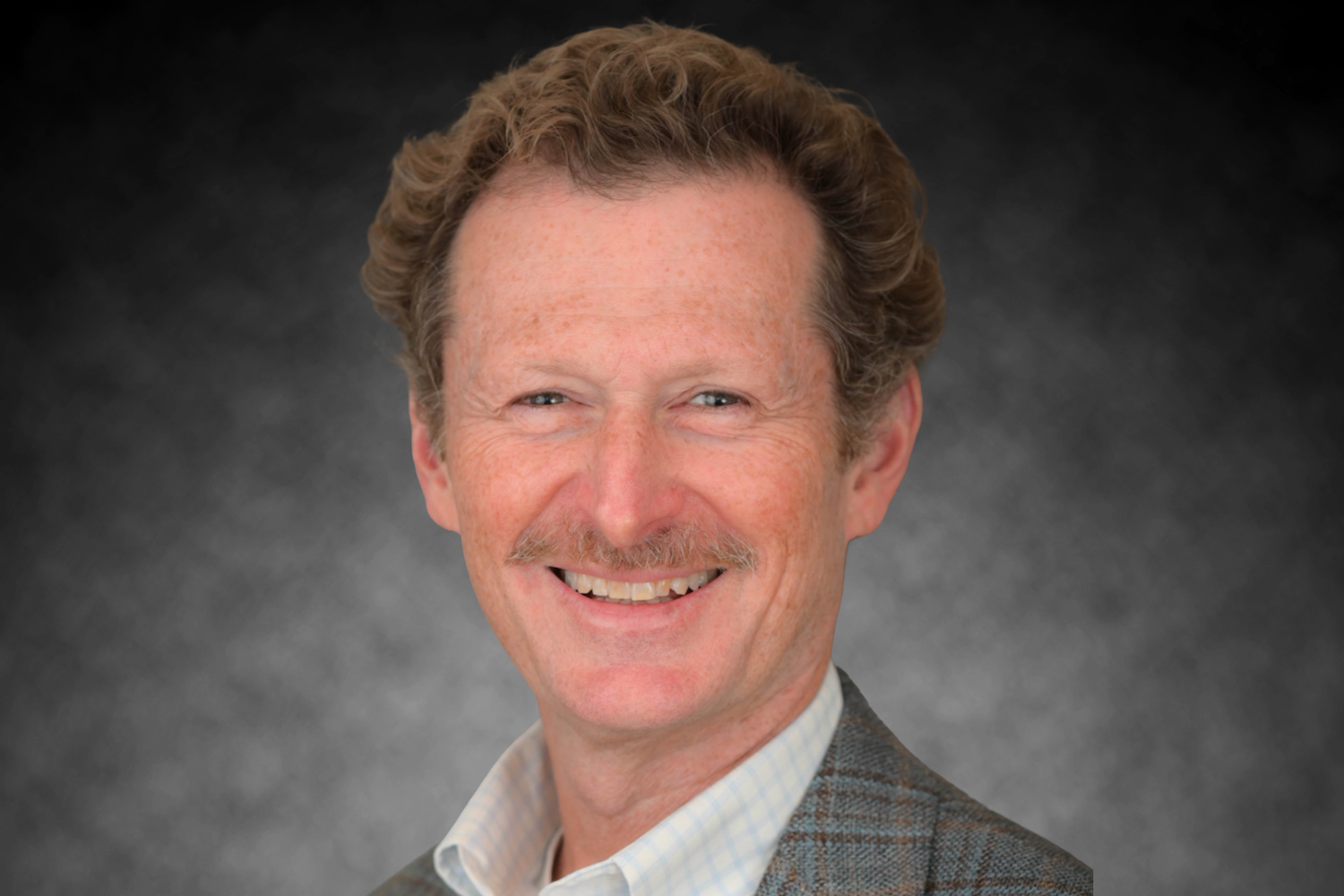 Smiling man with light brown curly hair and mustache, wearing a checkered blazer.