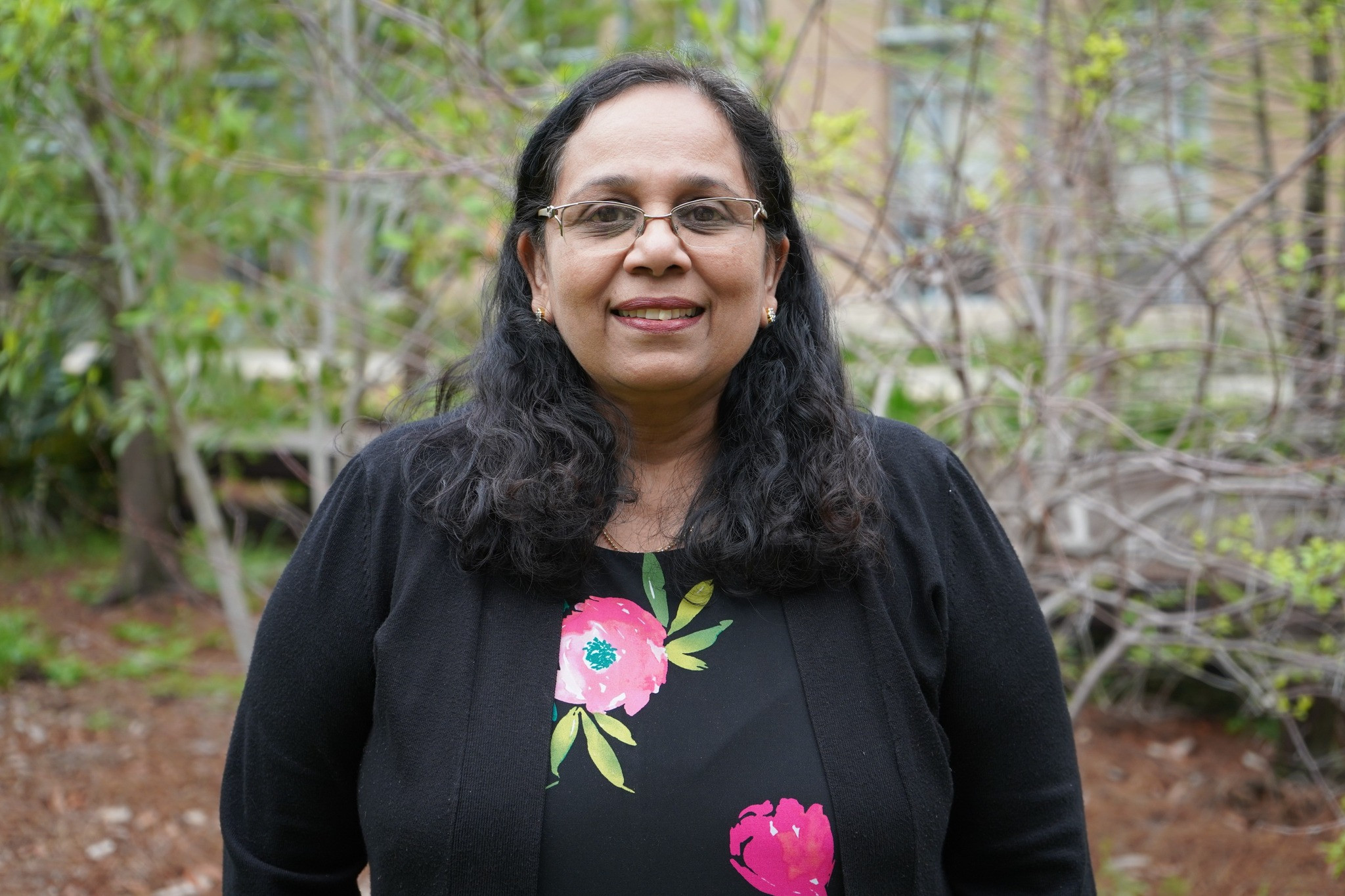 Smiling woman with dark hair and glasses wearing a black floral top, green foliage background.