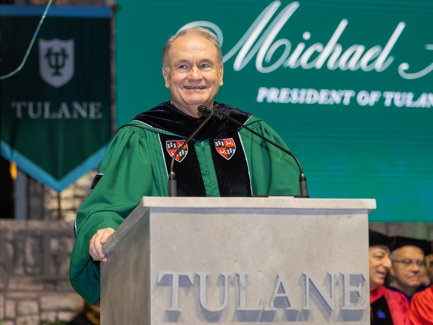 President Fitts green academic gown speaks at a podium with Tulane branding.