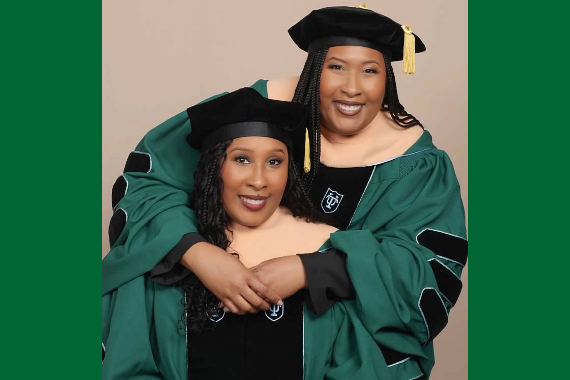 Two smiling women in green academic gowns and caps, one embracing the other.