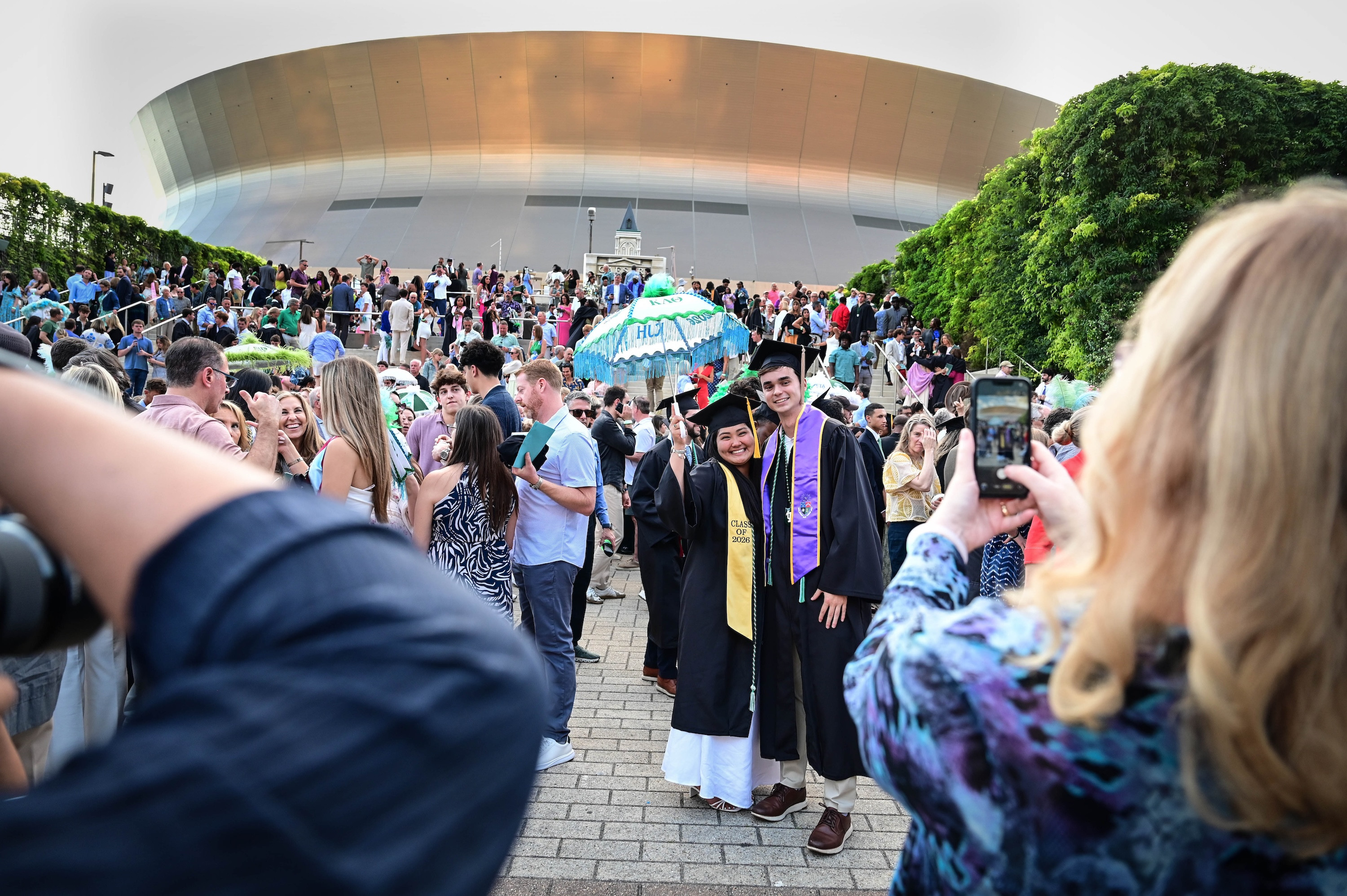 Two graduates in caps and gowns pose for a photo outside a gold-domed stadium.