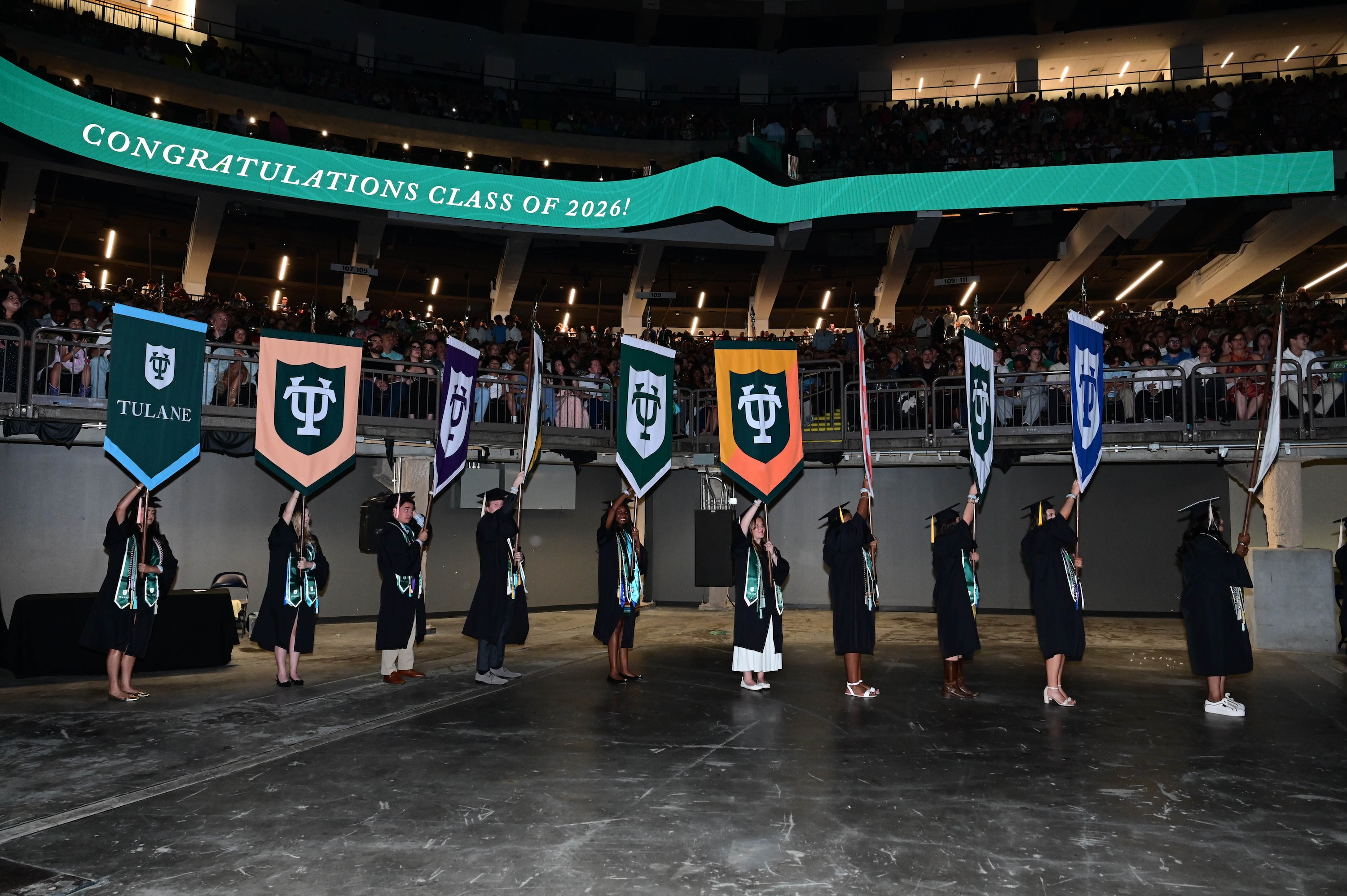 Graduates in caps and gowns hold colorful university gonfalons high in a large arena.