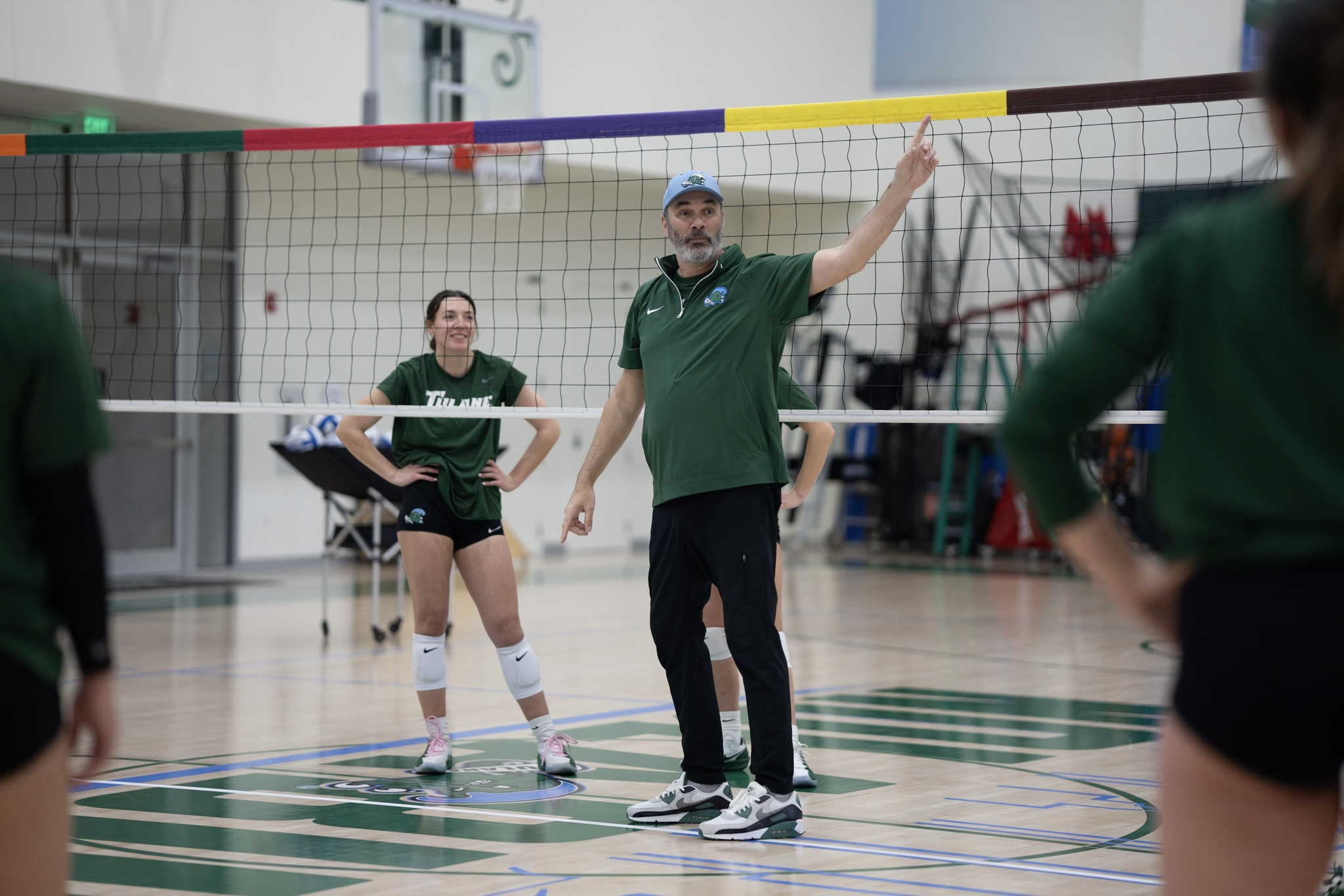 Volleyball coach in a green shirt gestures to female players on an indoor court.